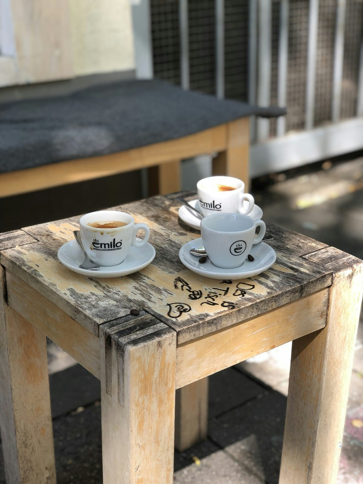 A close up of three white coffee cups and saucers on a small, worn wooden table with some graffiti on it. 