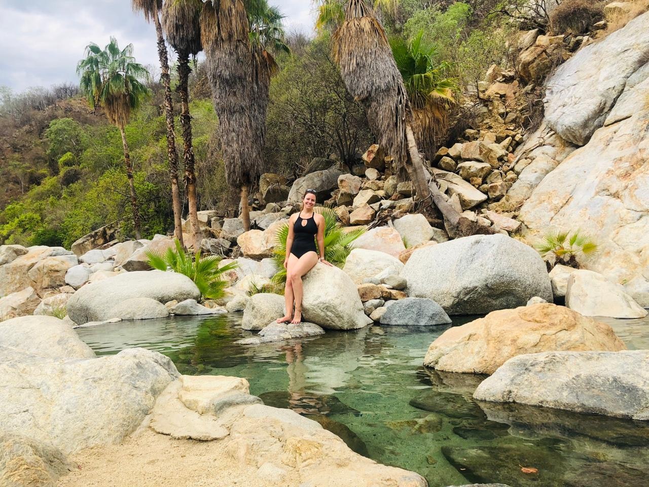 A woman in a black swimsuit poses against the rocks surrounding a hot spring.
