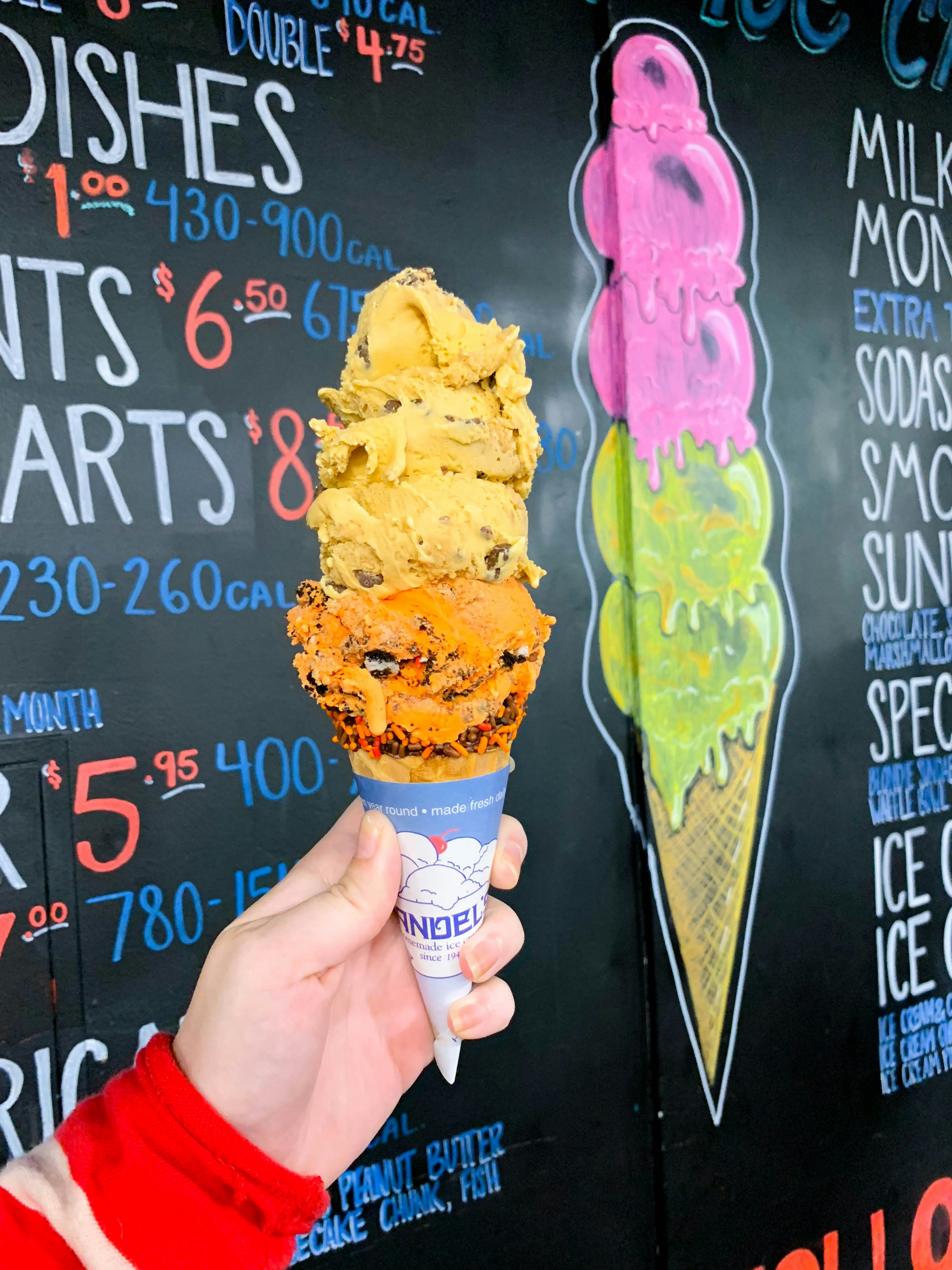 Four scoops of ice cream are stacked on a cone being held by a hand in front of a chalkboard offering other scoops; California ice cream