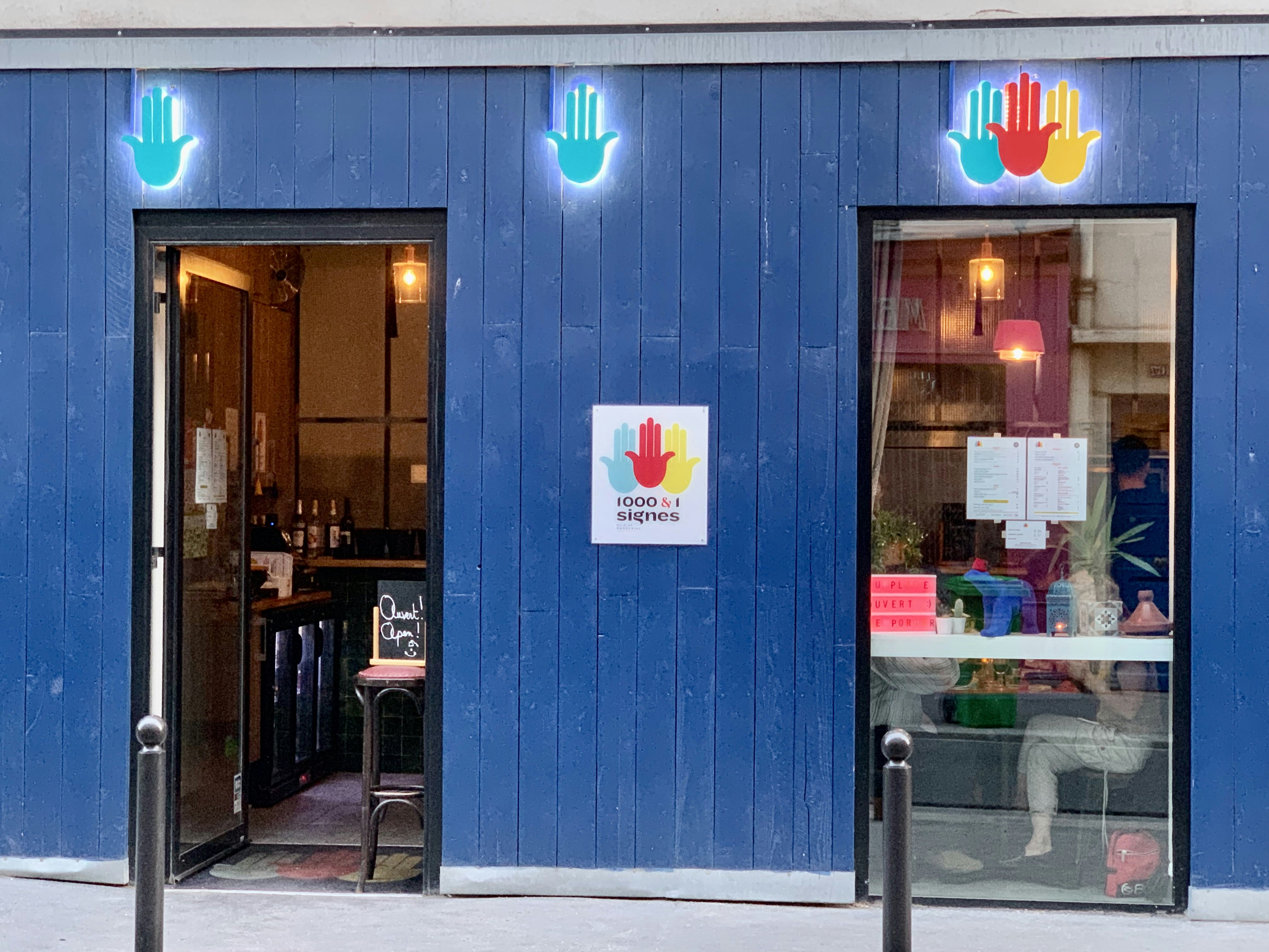 The cobalt blue entrance to 1000&1 Signes is marked by two blue hands in a Hamsa sign backlit with white neon and a cluster of three Hamsa hands in blue, red, and yellow over a tall window the same dimensions as the doorway to its immediate left. A diner can be seen faintly through the window