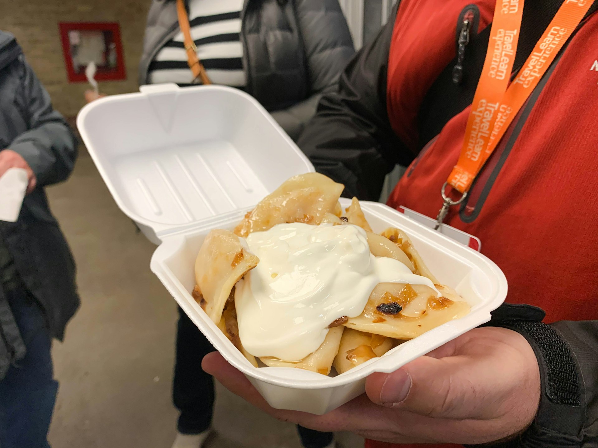 A hand holds a container of sour-cream topped pierogi. 