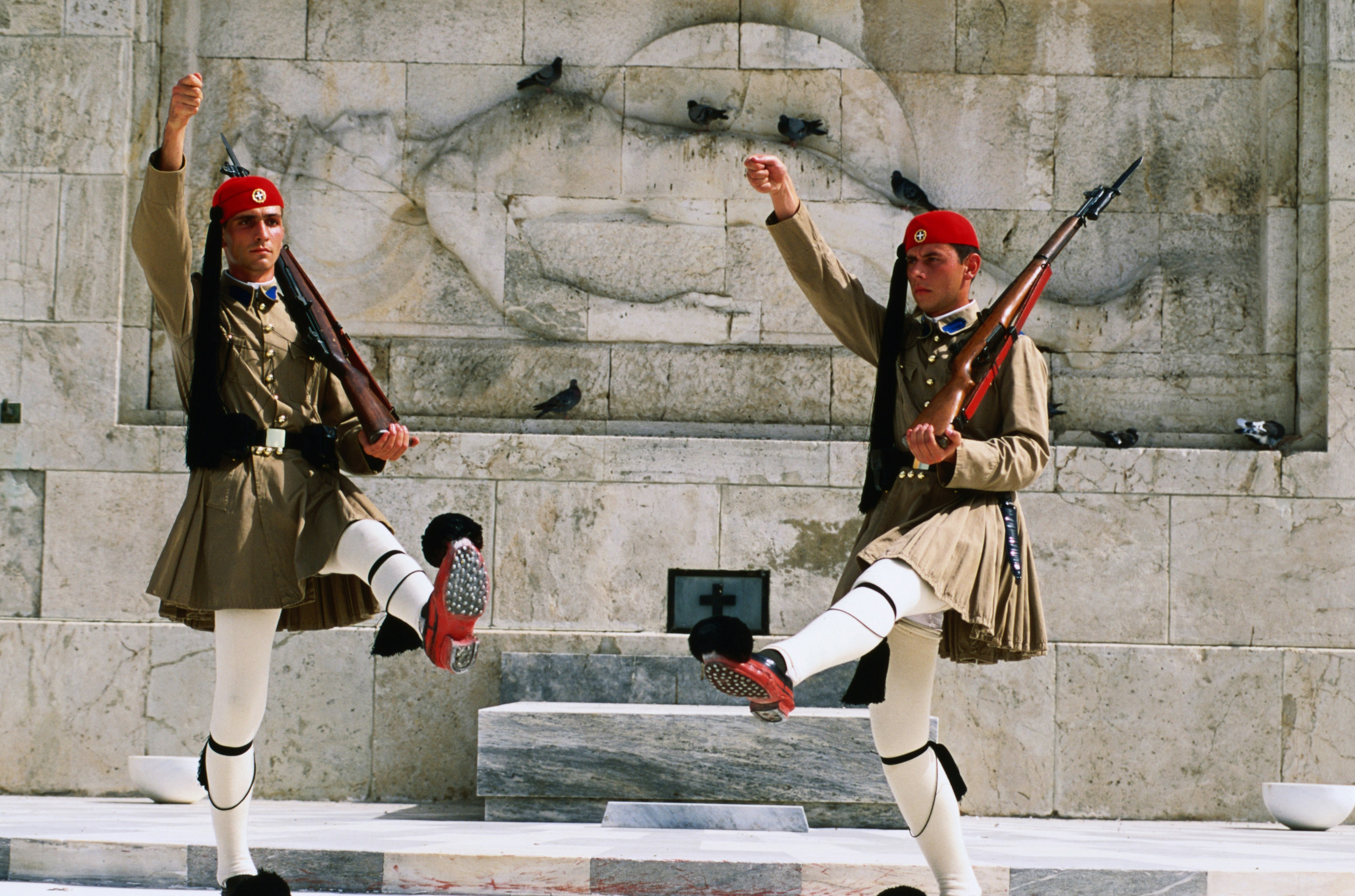 A pair of men wearing traditional garb with stockinged legs, a special kilt, fez hat and pom-pommed shoes each have an arm and a leg raised as they go though the traditional changing of the guard at the tomb of the unknown soldier in Athens