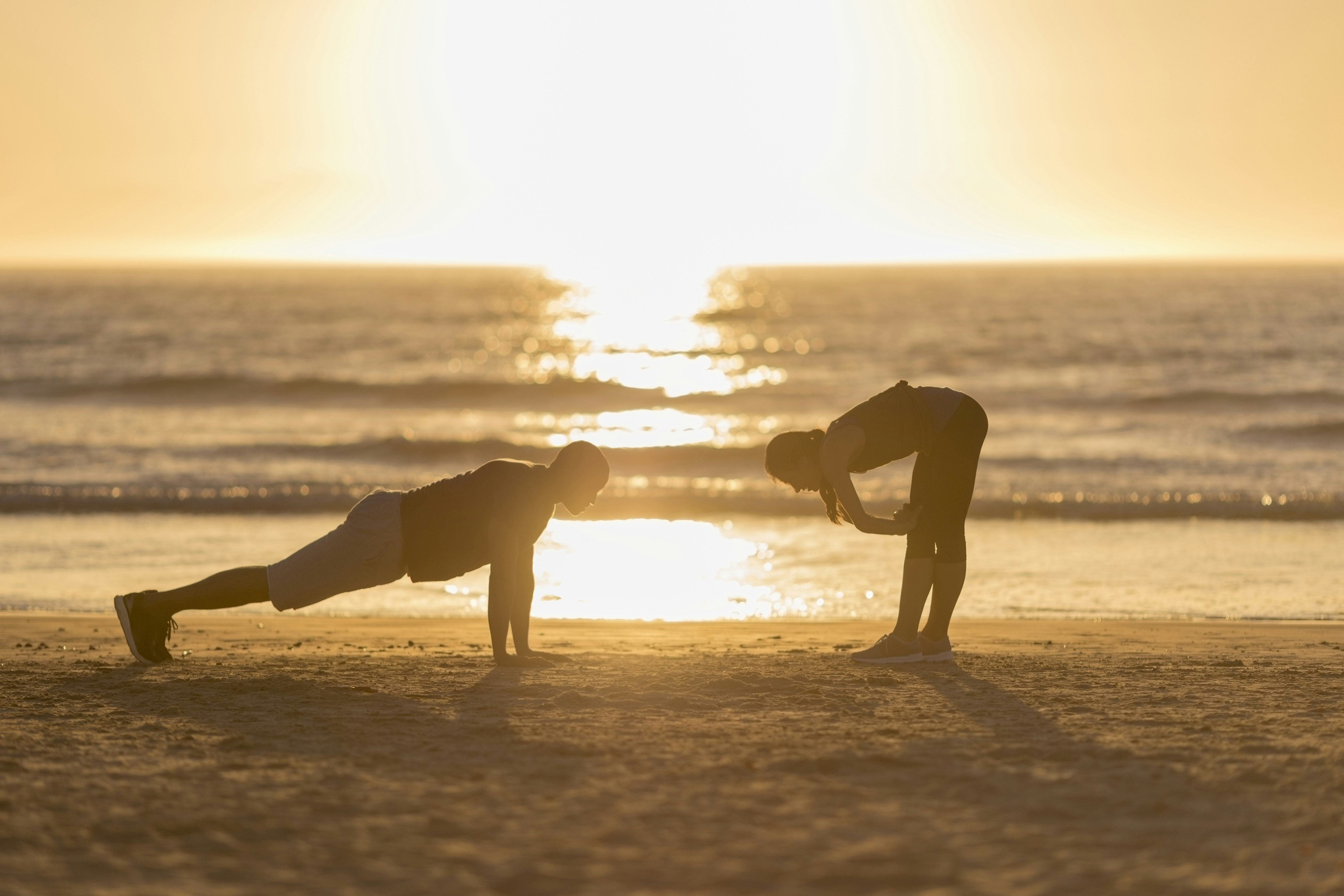 A man and woman exercise on a sandy beach at sunset. The male is mid-press up while the woman bends forward with her hands on her knees.