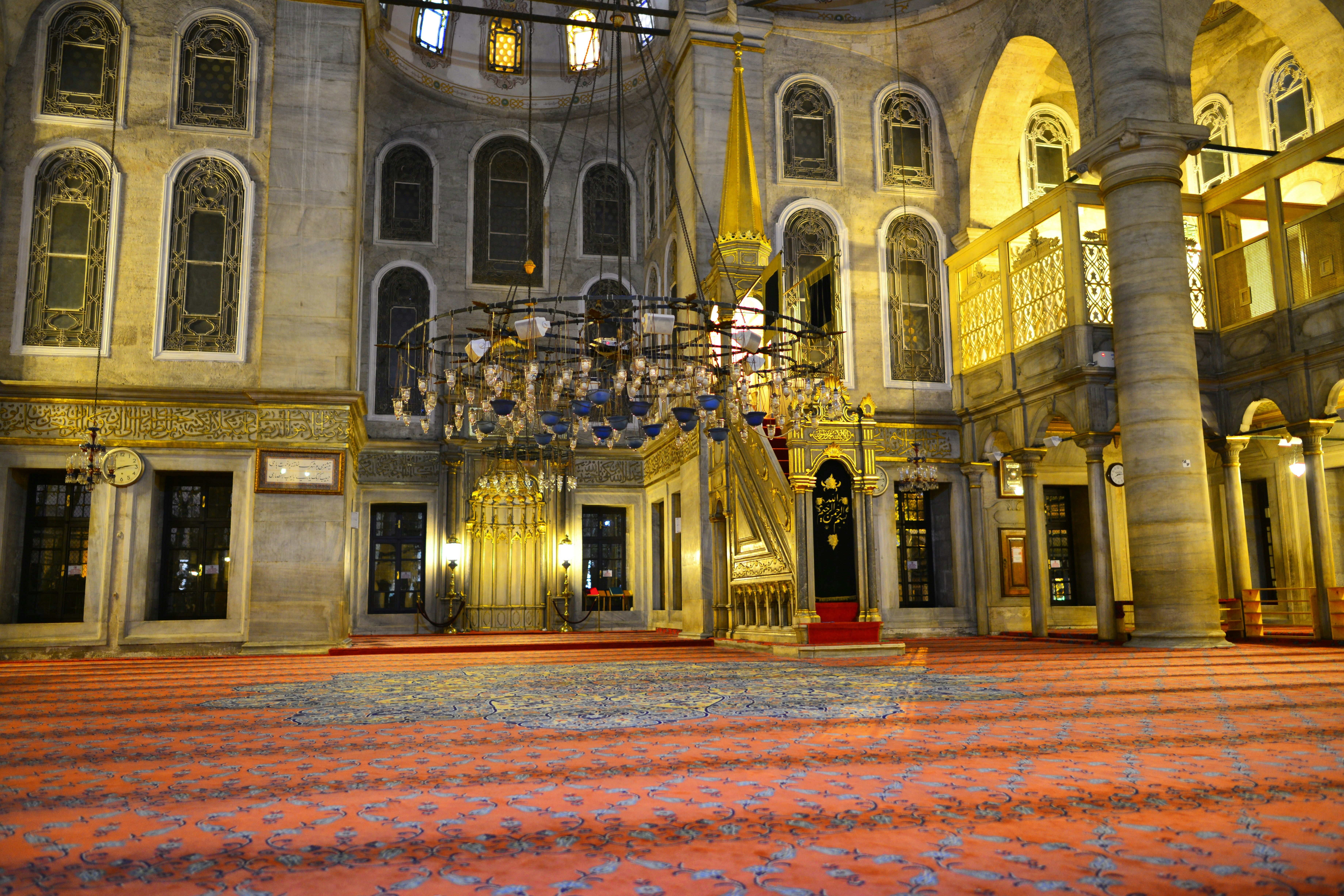 A large ornate chandelier hangs in the middle of the Eyüp Sultan Mosque. There's a large red and blue carpet spreading from one end of the mosque to the other.