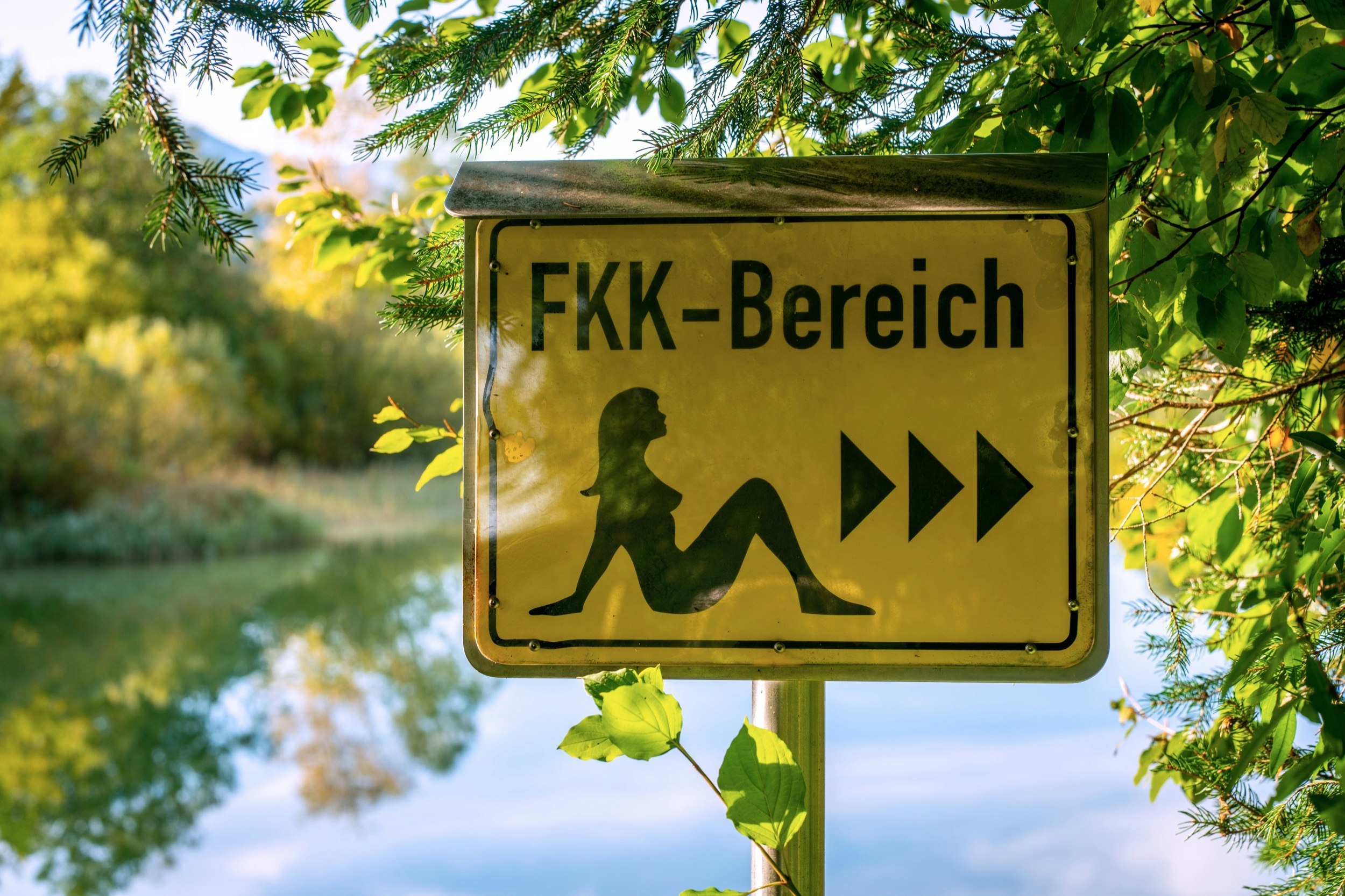A black and yellow sign with a figure of a naked lady sitting down and arrows pointing to the right with the words "FKK Bereich" is posted near a body of water.
