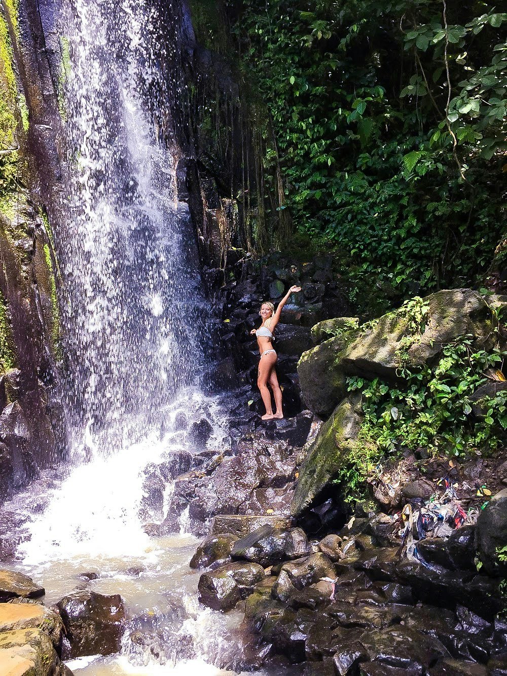 Fabiana stands beneath a waterfall in a white swimsuit.