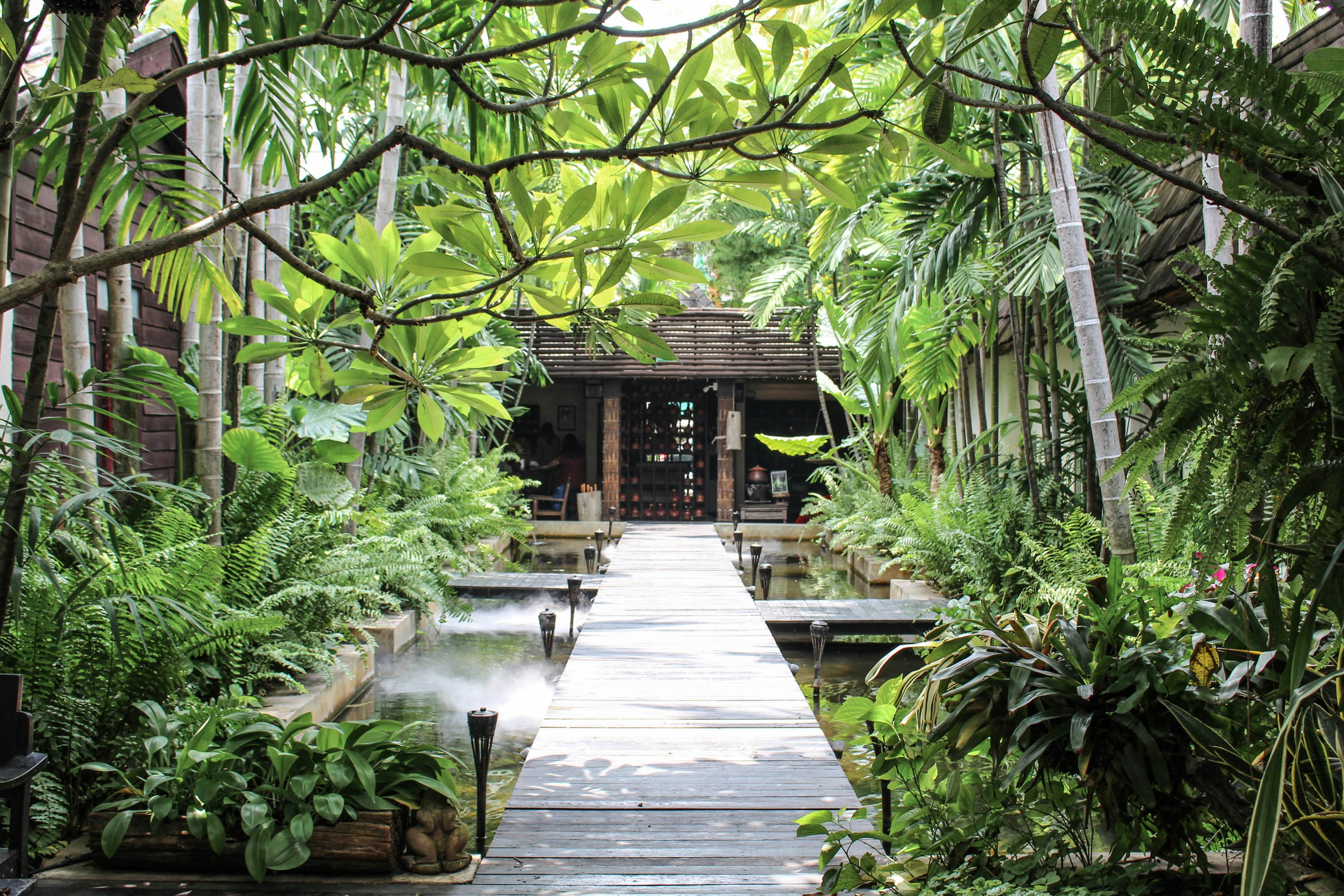 The entrance to Fah Lanna Spa in Chiang Mai: a narrow wooden boardwalk runs across a pond to the entrance of the facility, which is surrounded by greenery.