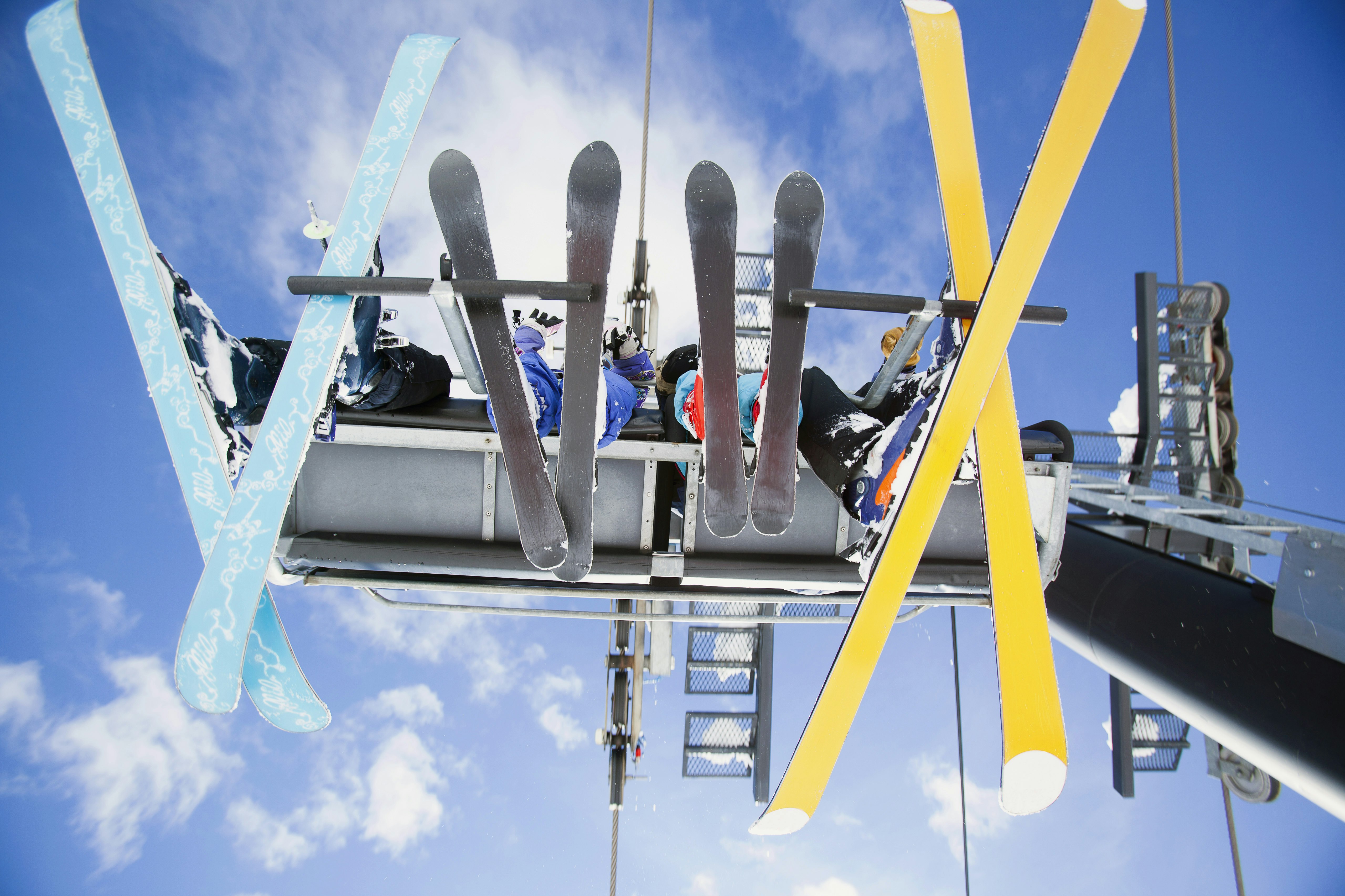 A family of four sit on a chairlift with their skis dangling below them. The image is taken from ground level looking up, with a cloudy blue sky providing the backdrop.