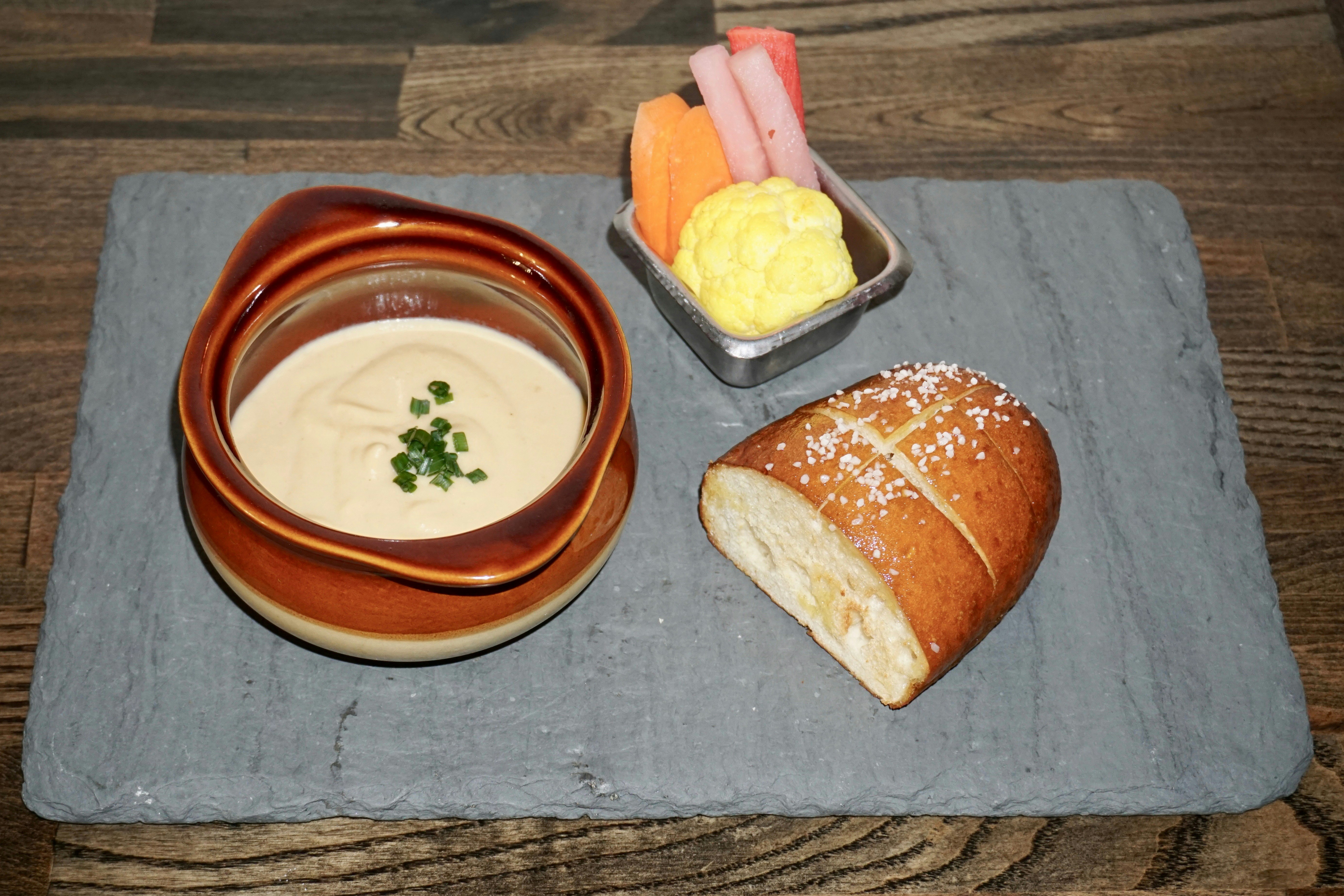 A gray slate, being used as a plate, sits on a dark wooden table; on the slate is a chunk of golden brown bread, a metal ramekin filled with crudités, and an earthenware bowl filled with molten vegan cheese, garnished with green herbs.
