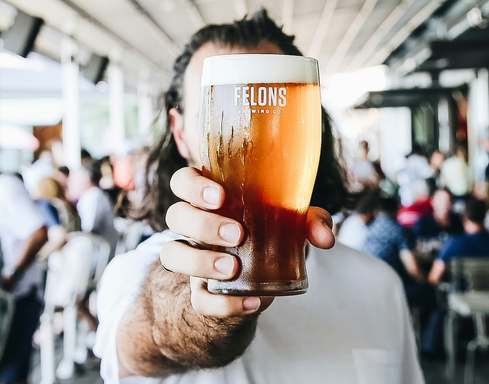 A man's outstretched hand holds a pint of beer up to the camera lens, obscuring his face behind the glass.