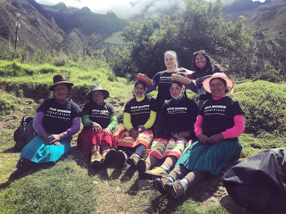 A group of female porters in Peru pose for the camera on a grassy hill along the Inca Trail. They are wearing black shirts that say 'Wild Women Expeditions' over their long skirts and long-sleeve shirts 