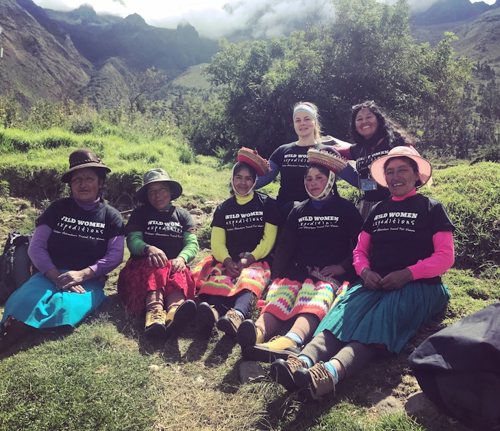 A group of female porters in Peru pose for the camera on a grassy hill along the Inca Trail. They are wearing black shirts that say 'Wild Women Expeditions' over their long skirts and long-sleeve shirts 