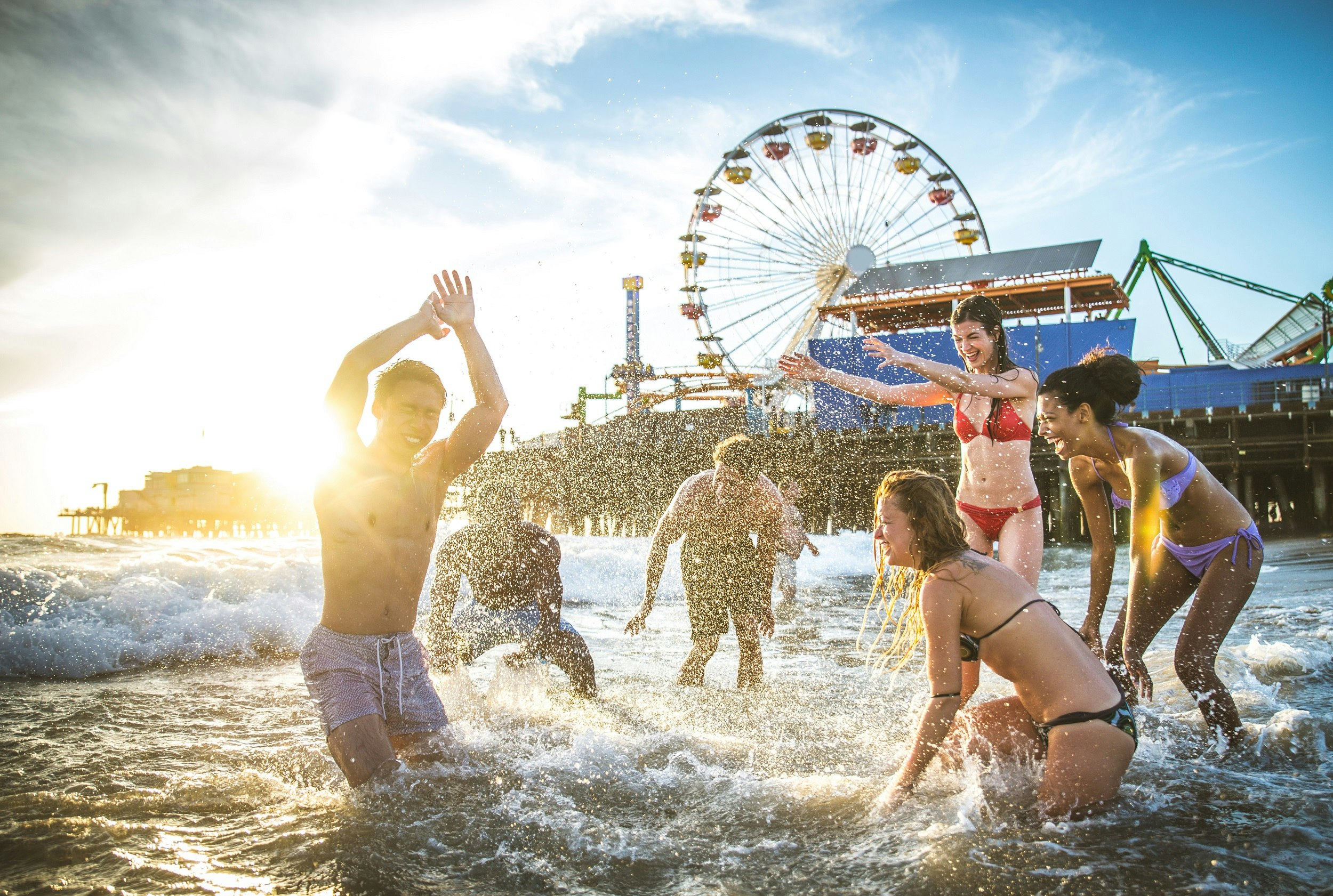 A group of young adults play in the surf, with the Santa Monica Pier ferris wheel in the background.