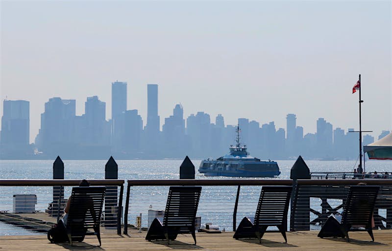 A woman sits in a reclining lounge chair next to a railing overlooking a waterway in Vancouver. A ferry sails through the waterway toward the far shore which is dominated by skyscrapers.