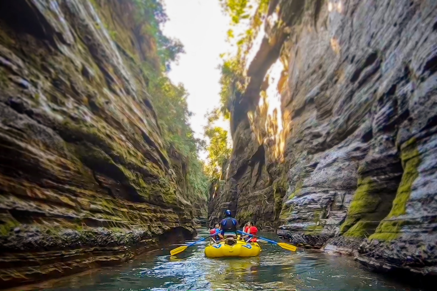 A yellow inflated raft filled with people wearing helmets, life jackets and carrying paddles, travels down a waterway along side two massive rock formations