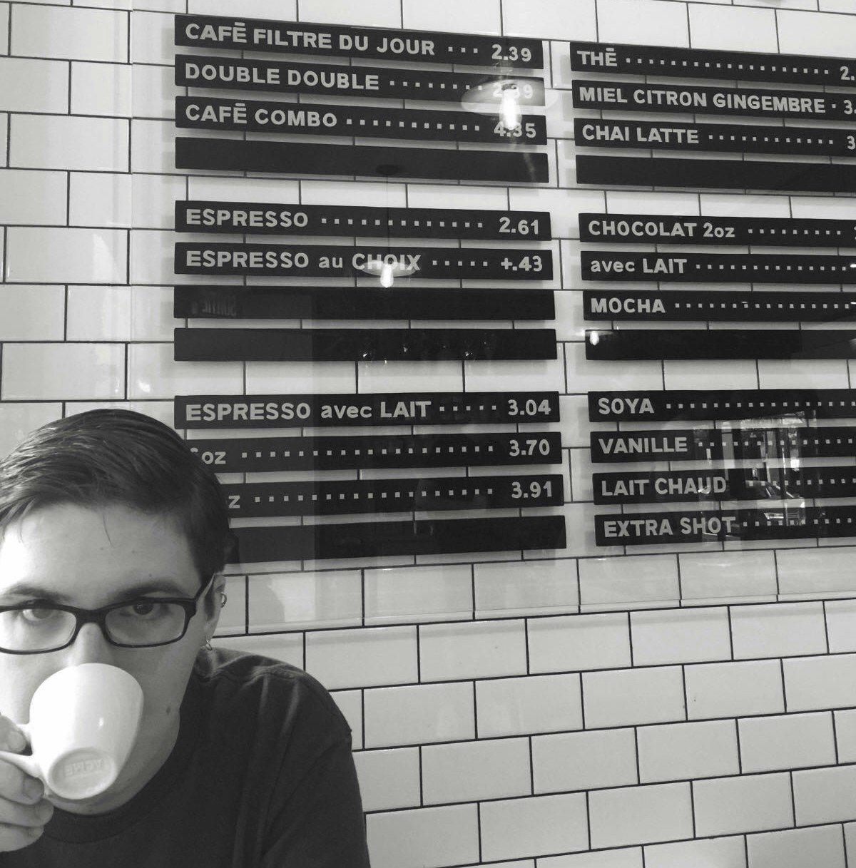 Article writer Fionn Pooler sits under a coffee shop price list mounted on a white tiled wall. He's looks directly at the camera as he takes a sip of coffee.