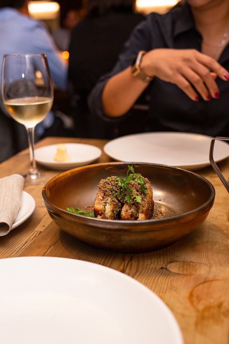 A brown ceramic bowl sitting in the middle of a table, with a freshly prepared dish in it, which is garnished with fresh herbs. A diner sitting at the place setting opposite is in soft focus.