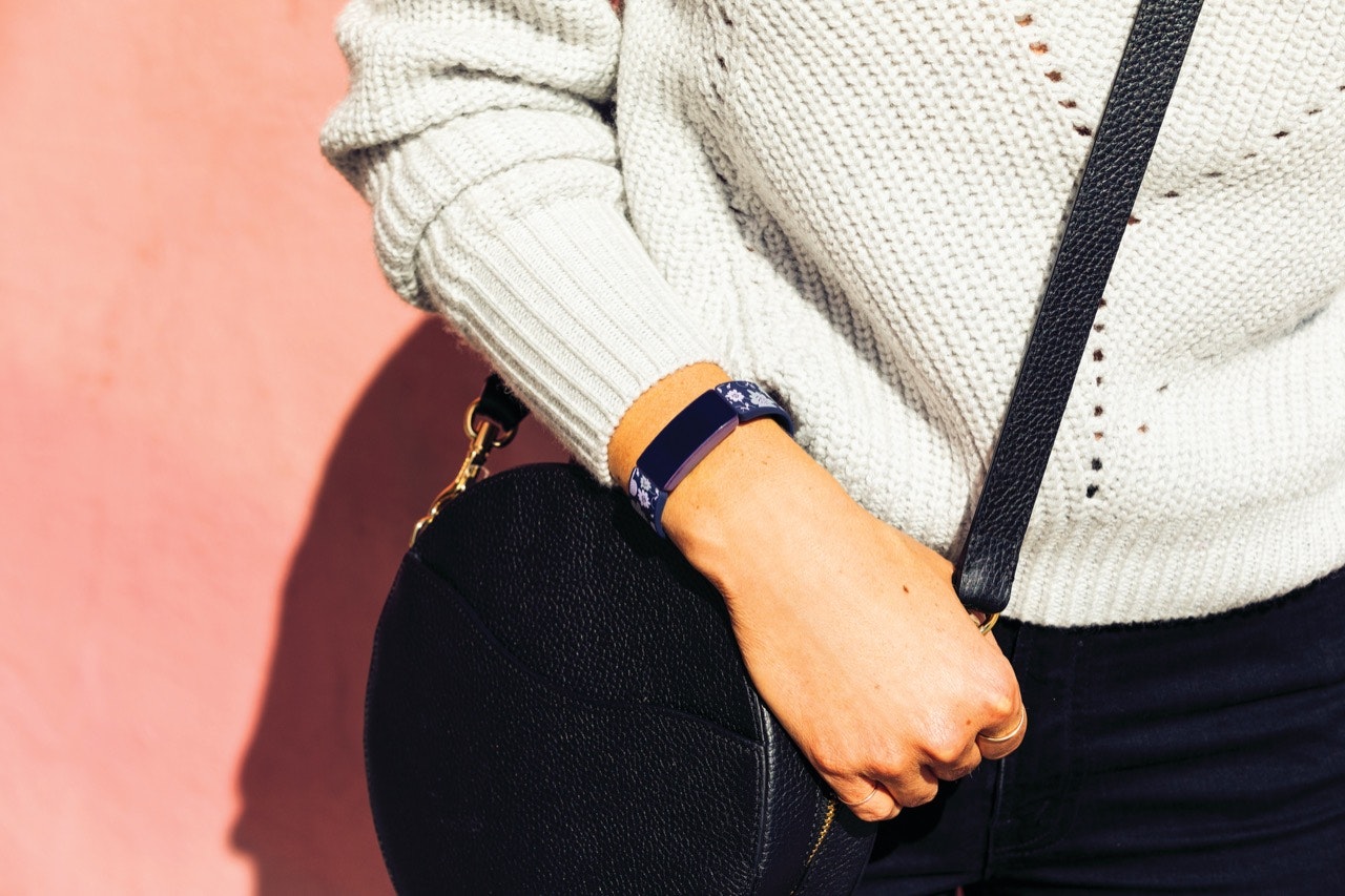 Closeup on person wearing a white sweater and a circular black crossbody bag, holding the strap with Fitbit's Inspire HR on their wrist