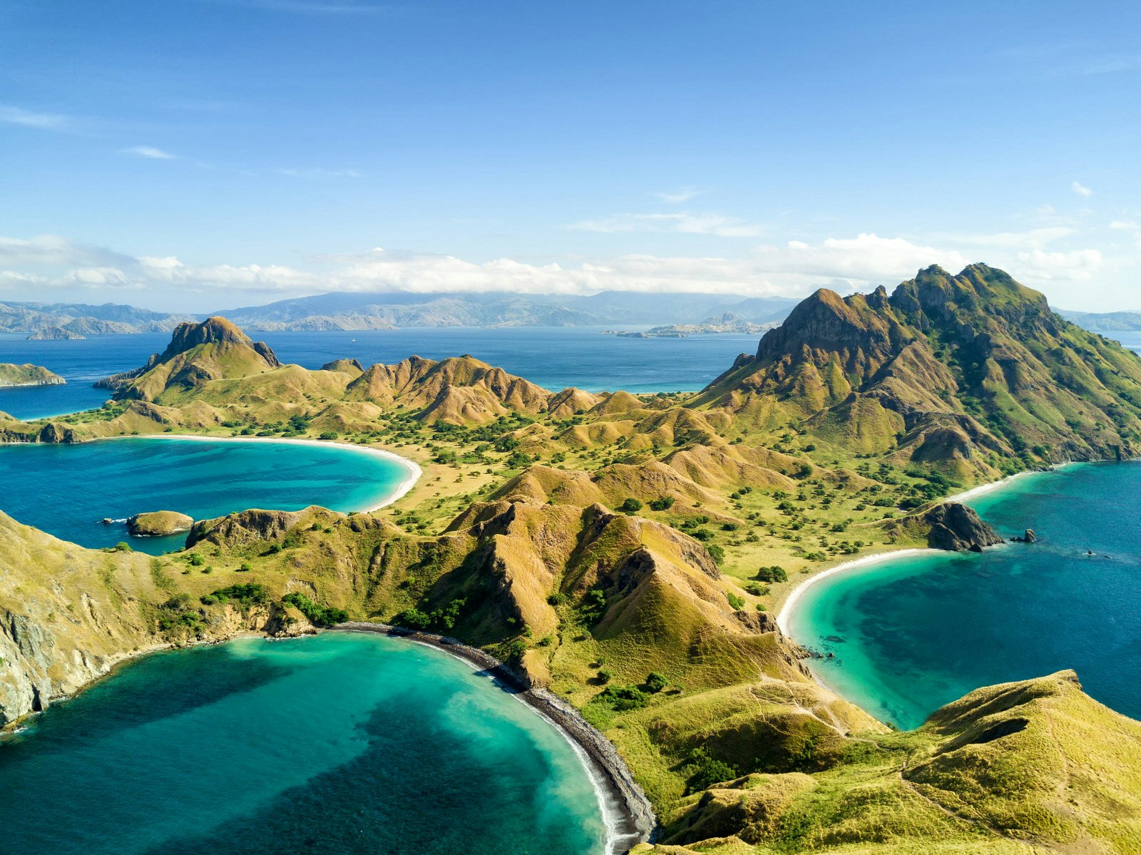 Aerial of the mountainous Pulau Padar island, Indonesia. Taken on a sunny day with a few fluffy clouds in the far distance, the verdant green hills contrast against the deep, jewel-blue sea. 
