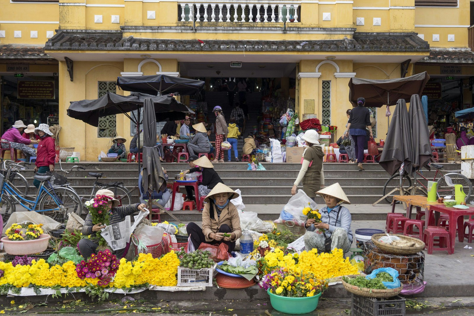Flower vendors on the concrete steps outside Hoi An central market. The women, wearing conical hats, are selling yellow and pink blooms; there are bikes and people in the background. 