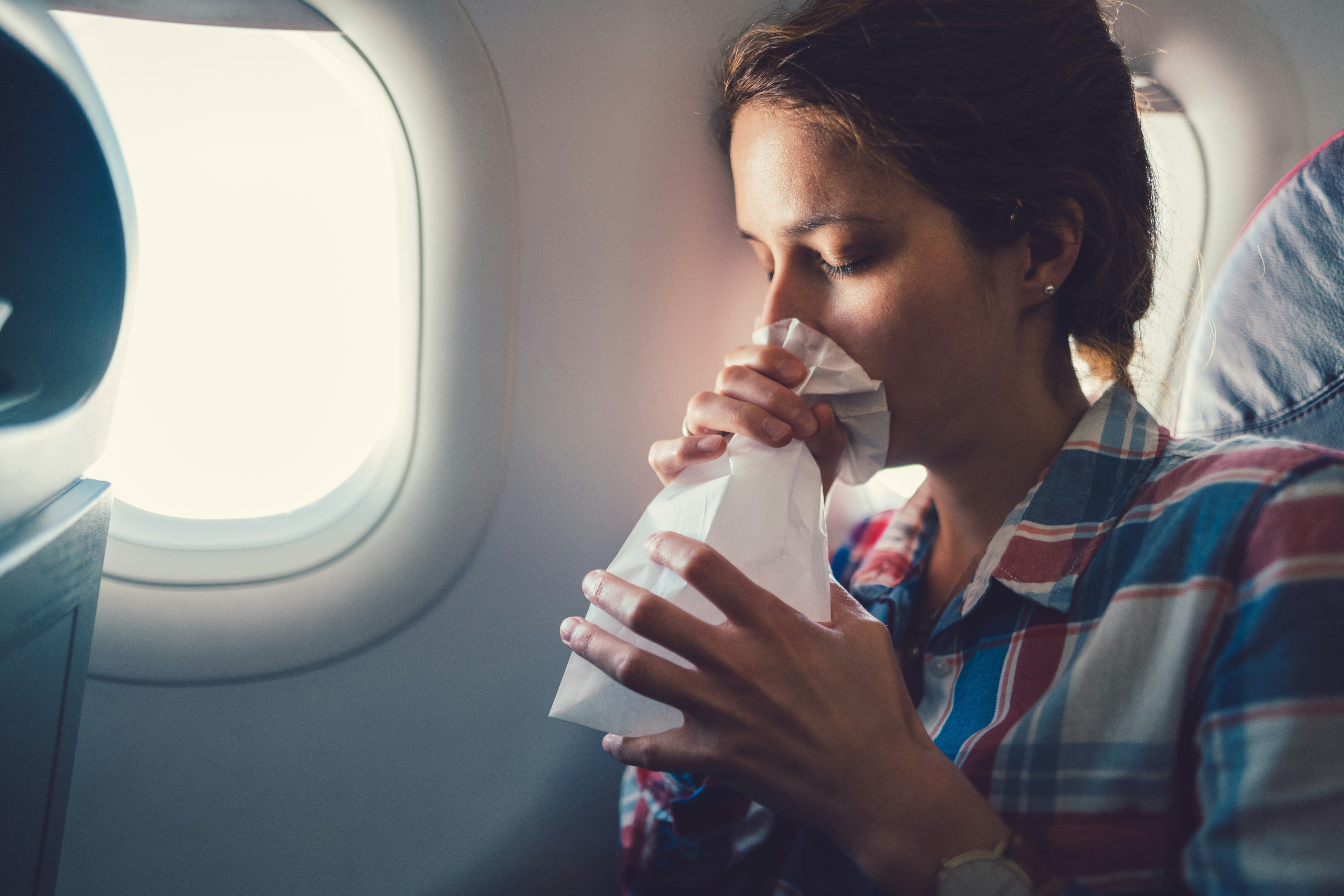 A woman sitting in a window seat on an airplane breathes into a white paper bag