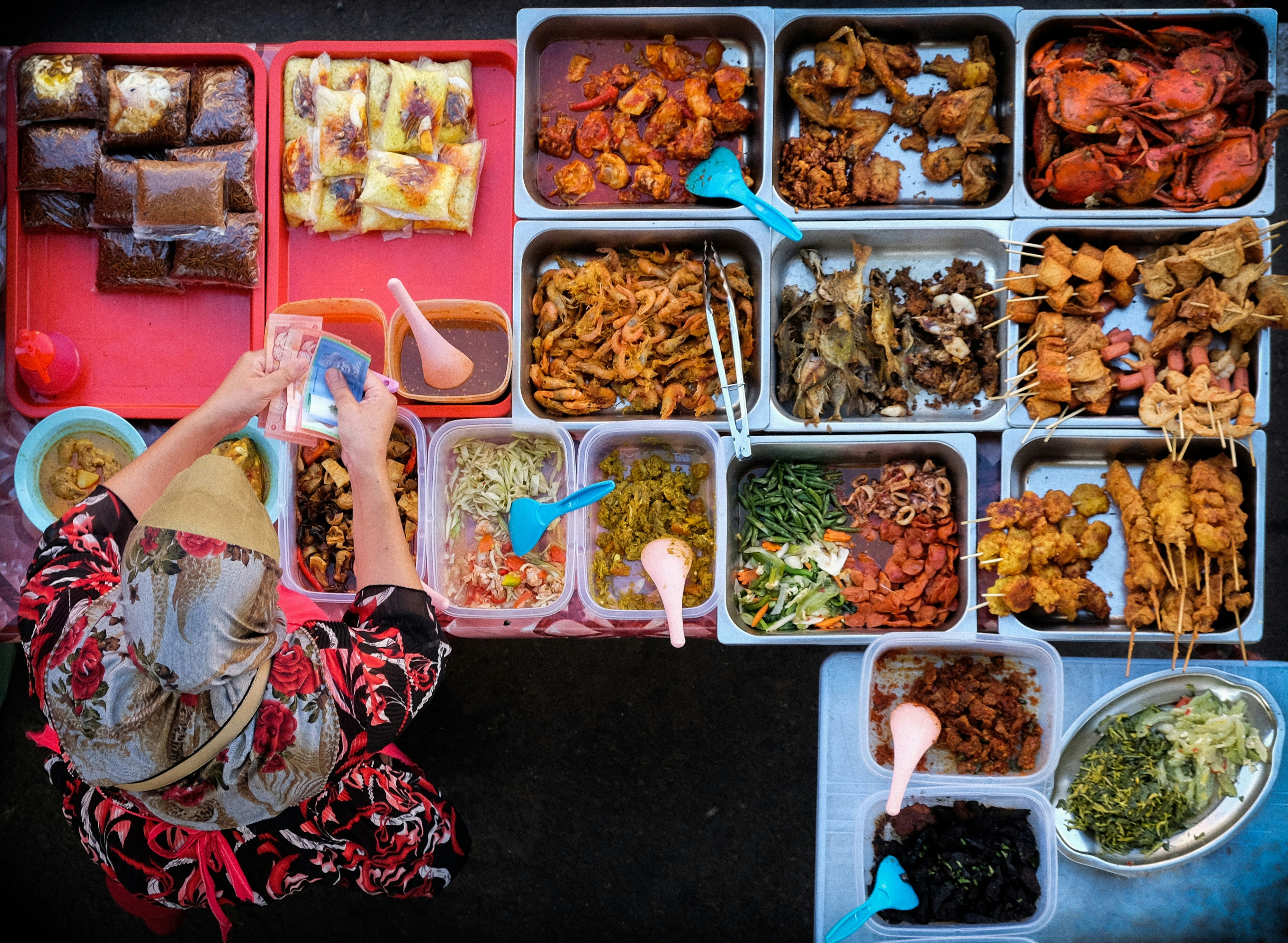 A top-down shot of a woman counting money at a food stall. The stall is filled with trays of colourful, Asian-style cuisine.