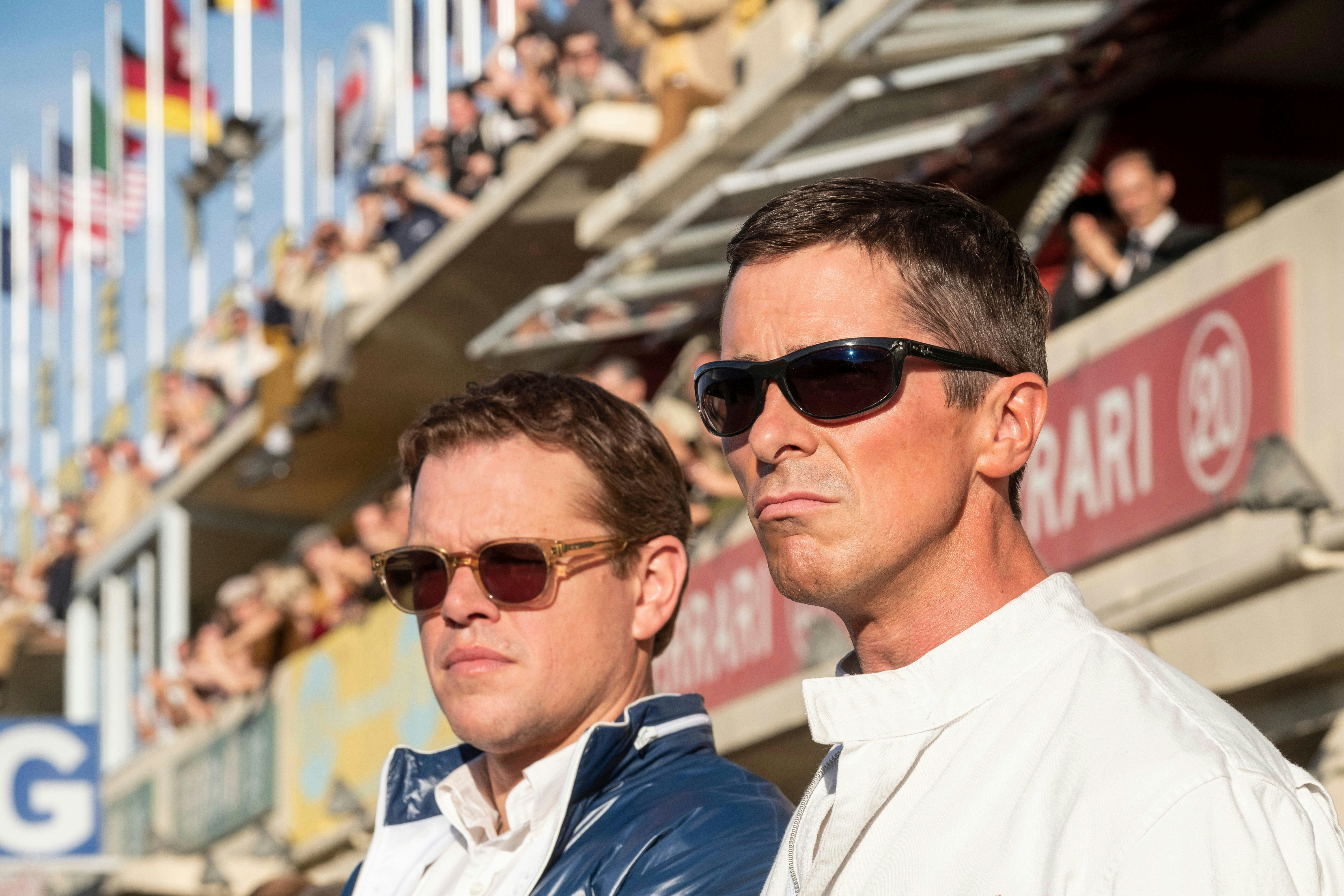 In a scene from the movie 'Ford v Ferrari', Matt Damon and Christian Bale stand side-by-side, looking out of frame, with stands filled with crowds behind them.