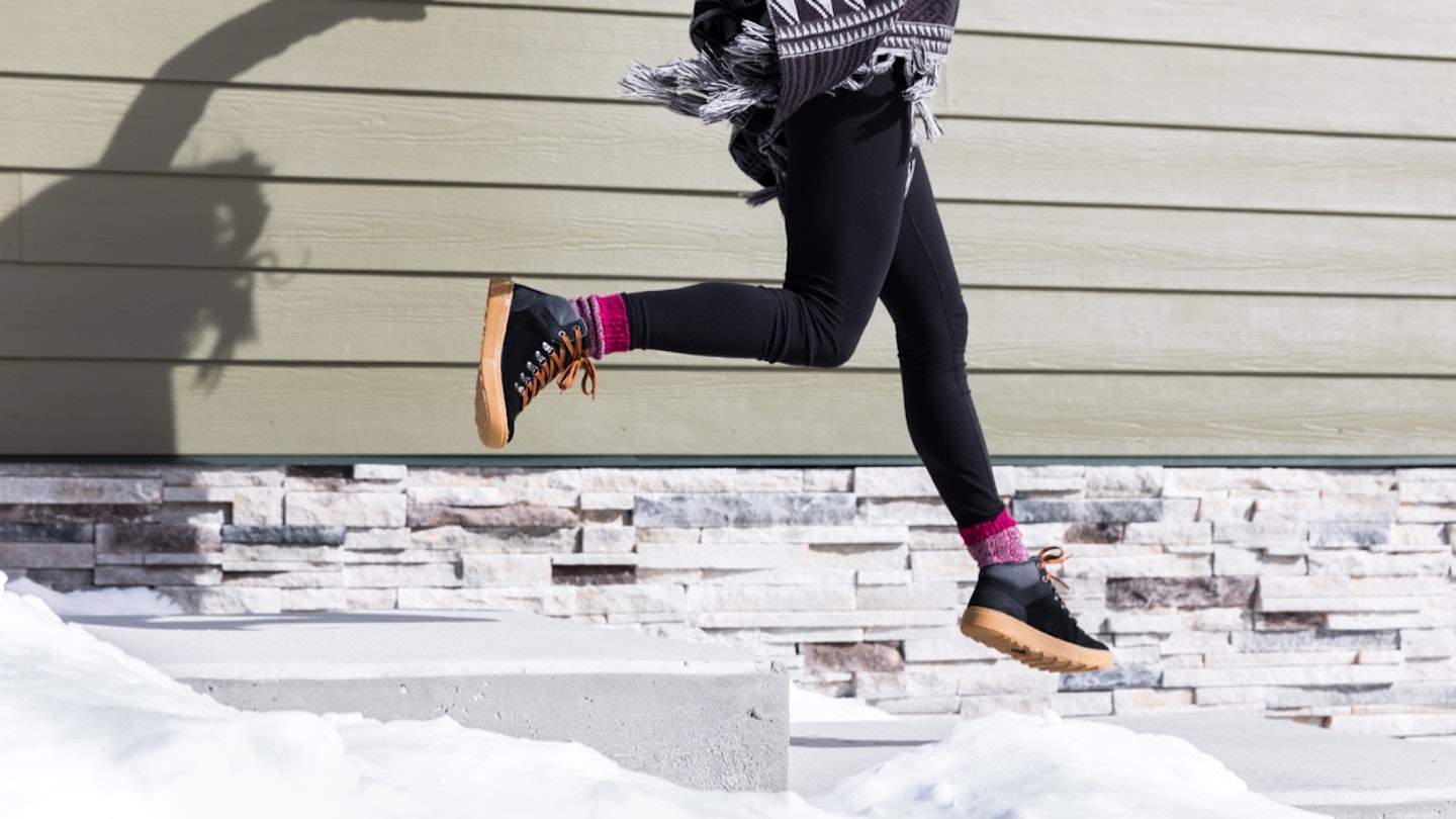 a woman jumping down snow-covered steps wearing Forsake's Lucie Mid boot in black