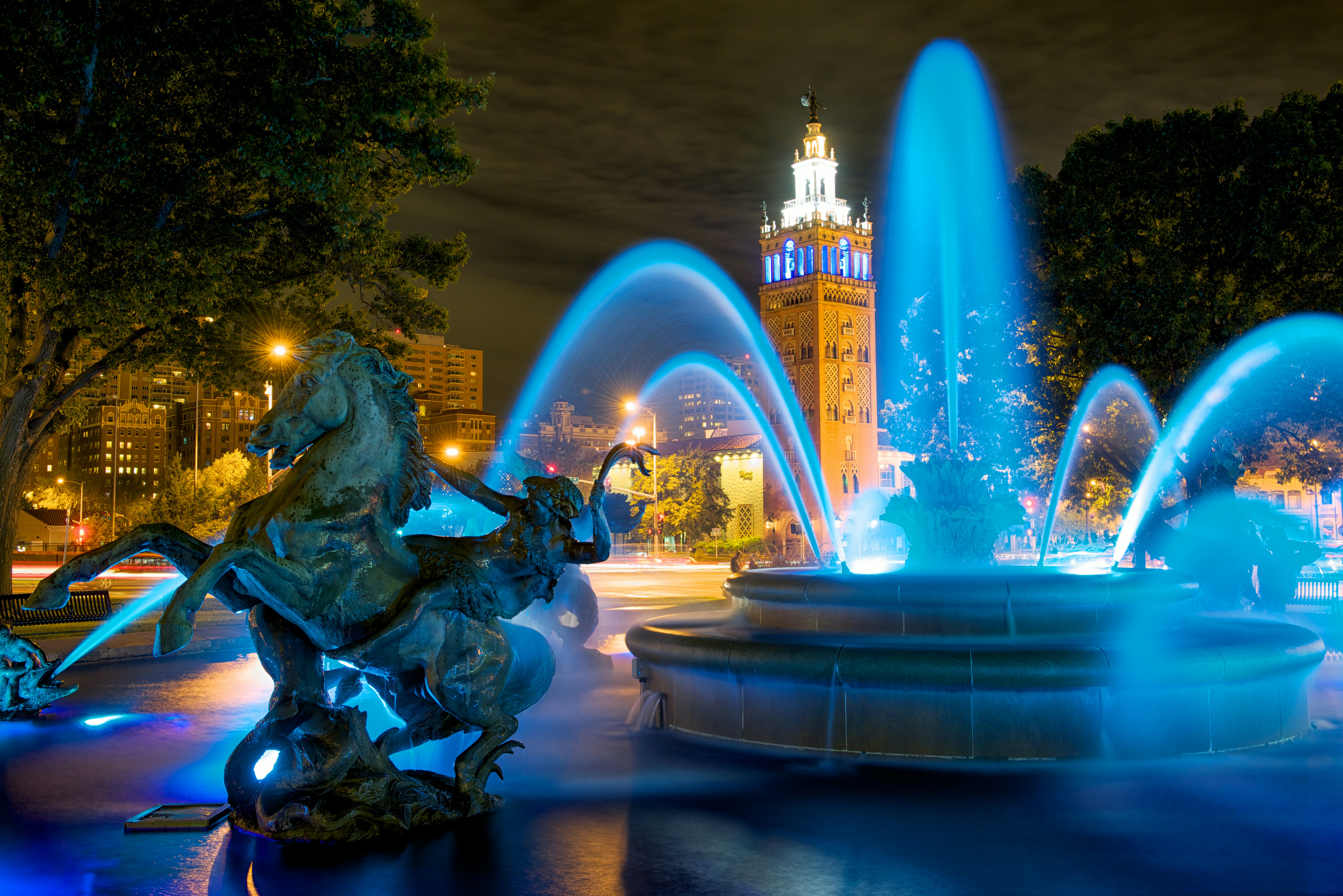 Streams of water is illuminated by blue light spray from a stone fountain. In front of the fountain is a man riding a large horse. 