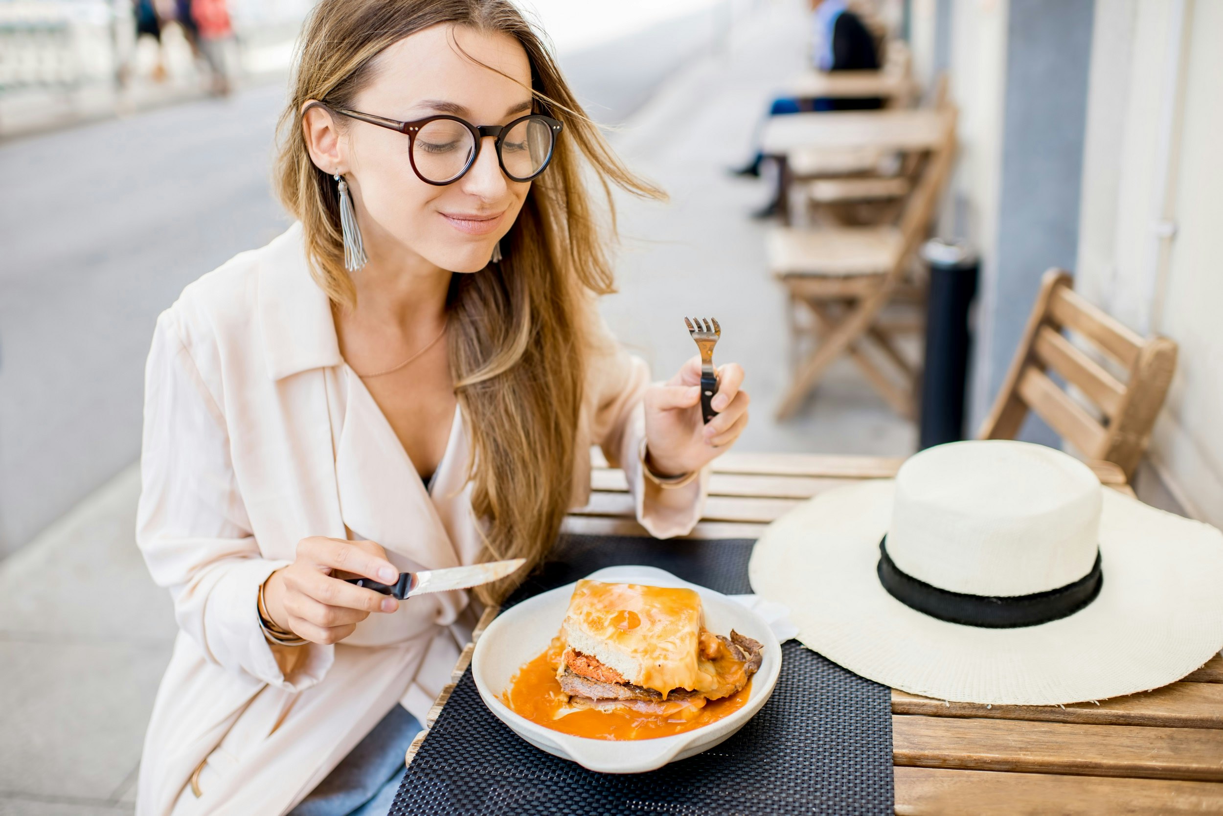 A woman with a full mouth, and wearing glasses, holds a knife and fork while gazing down at a large francesinha sandwich; it's topped with an egg and thick tomato sauce