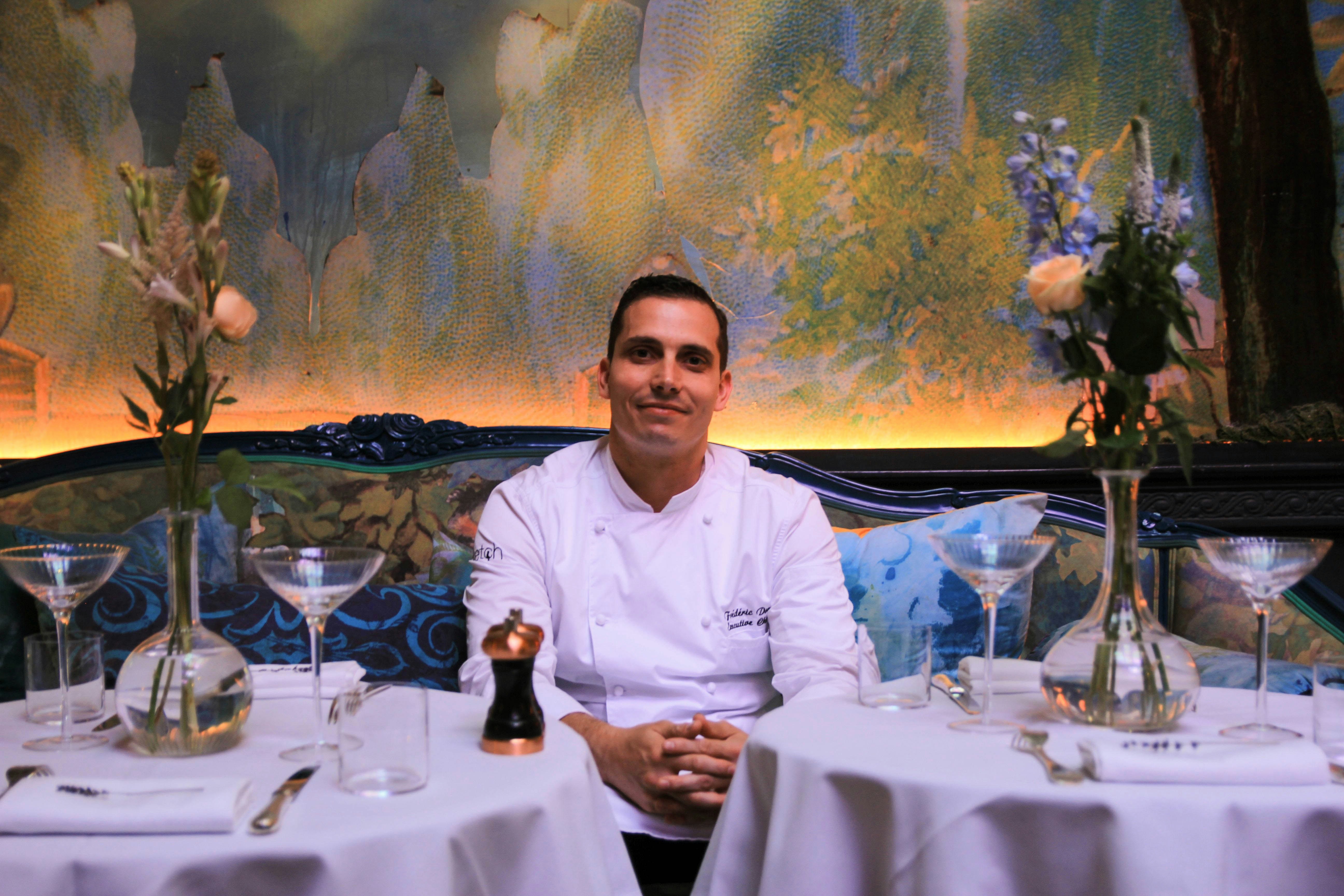 A chef in a white chef jacket sits between two round tables with white tablecloths, each set with a vase of flowers and wine glasses. The wall behind him is a painted mural in muted colours.