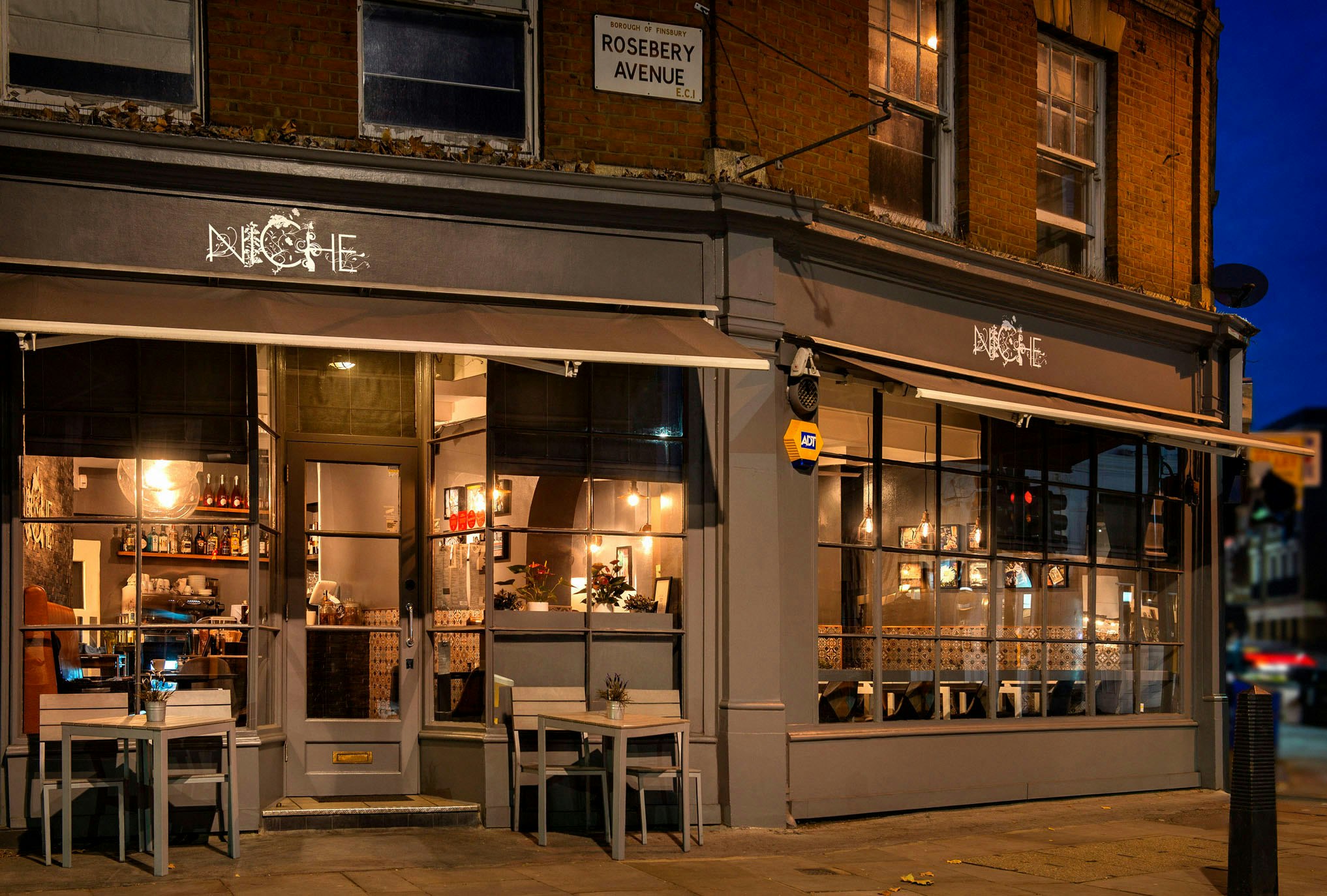Facade of a lit up restaurant on a London street corner with grey tables out front