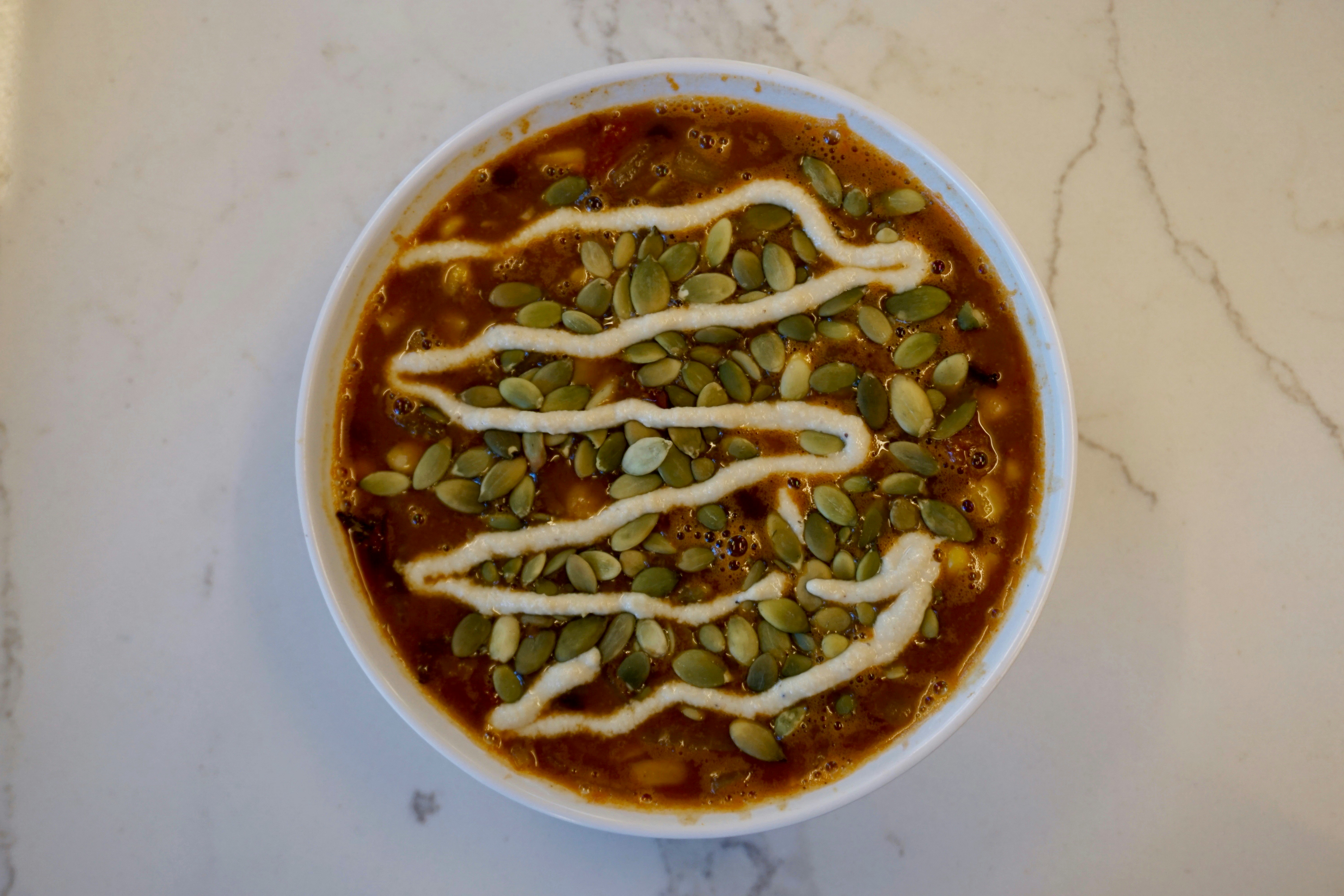 A white bowl sits on white marble counter; it's filled with a rich, reddish-brown, plant-based chili, which is topped with pumpkin seeds and a zig-zag of a creamy sauce.