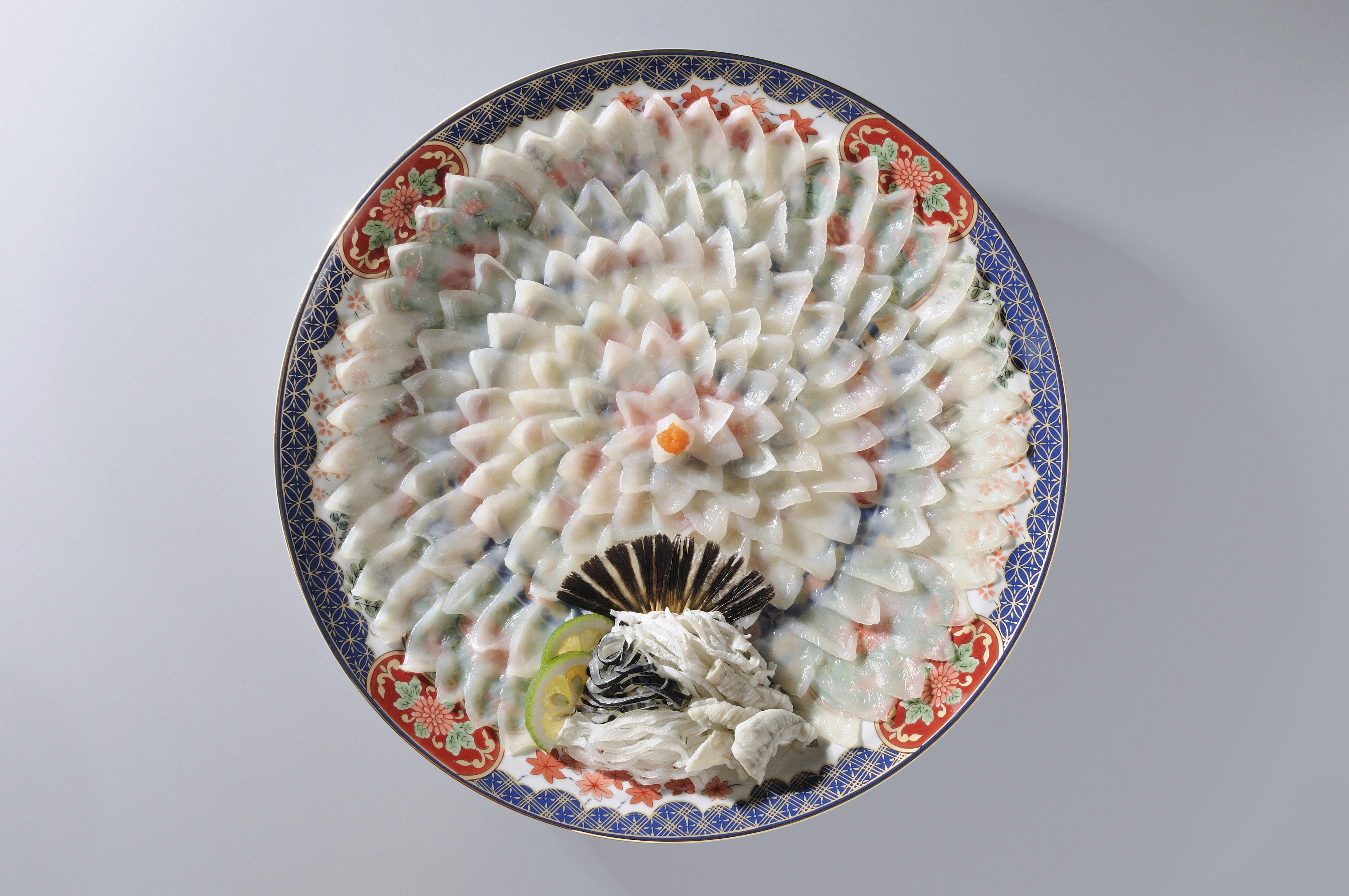 Thinly sliced fugu (puffer fish) is intricately arranged in the shape of a flower on a colorful plate. 