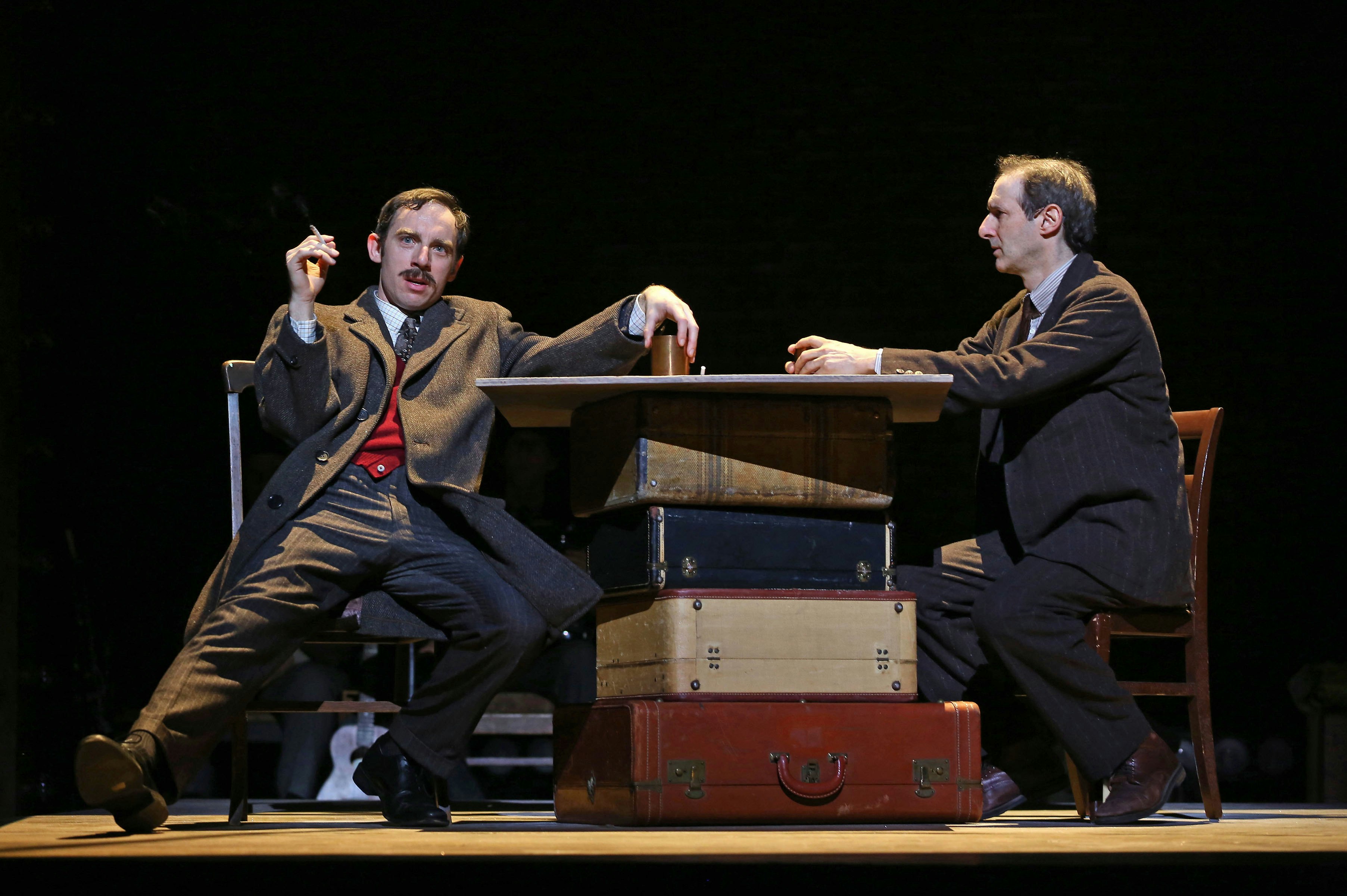 Two men sit at a table on a stage, one smoking.