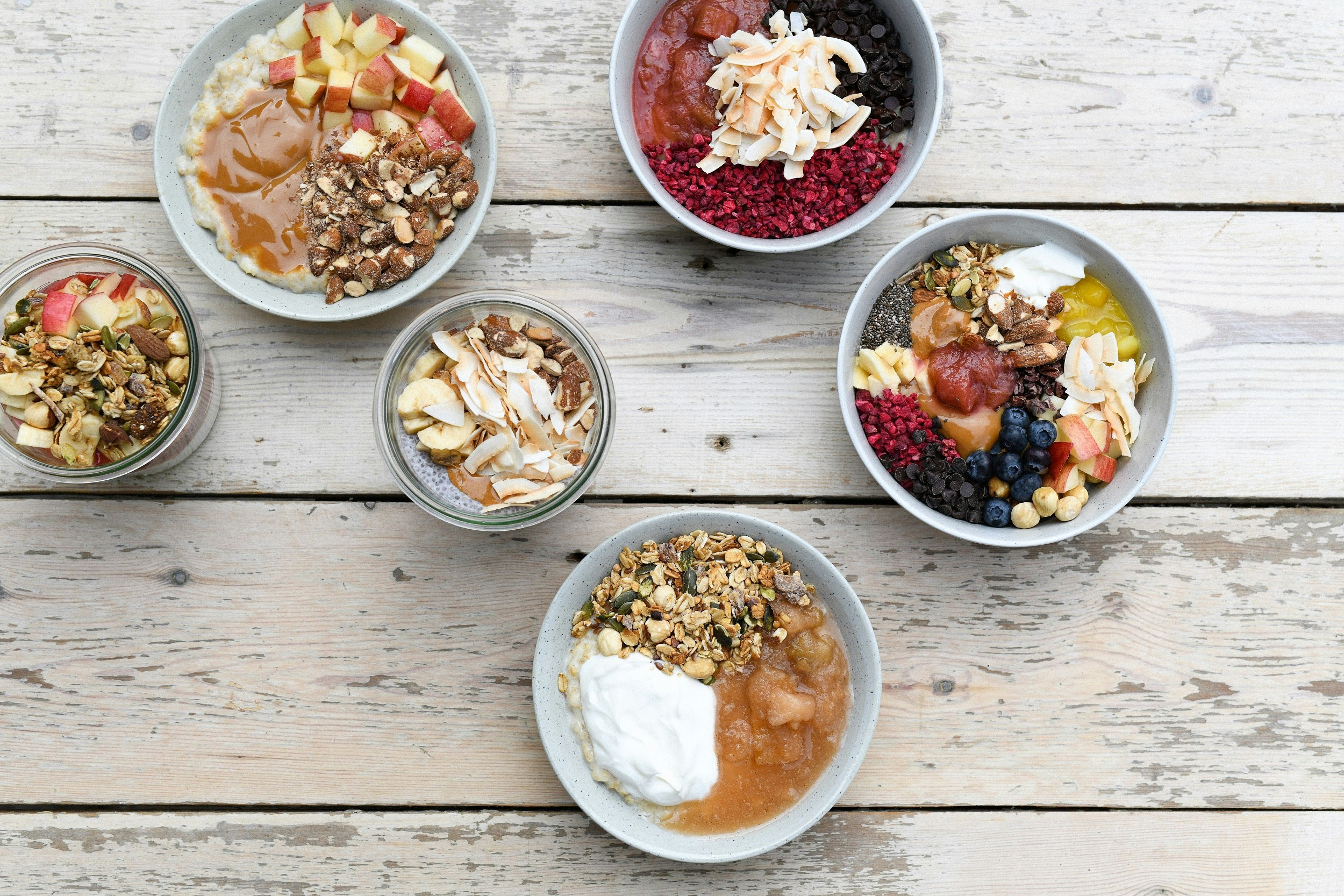 Four bowls and two glass jars sit on a white wooden table; each is full of different dishes, all being assortments of fruit and nuts.
