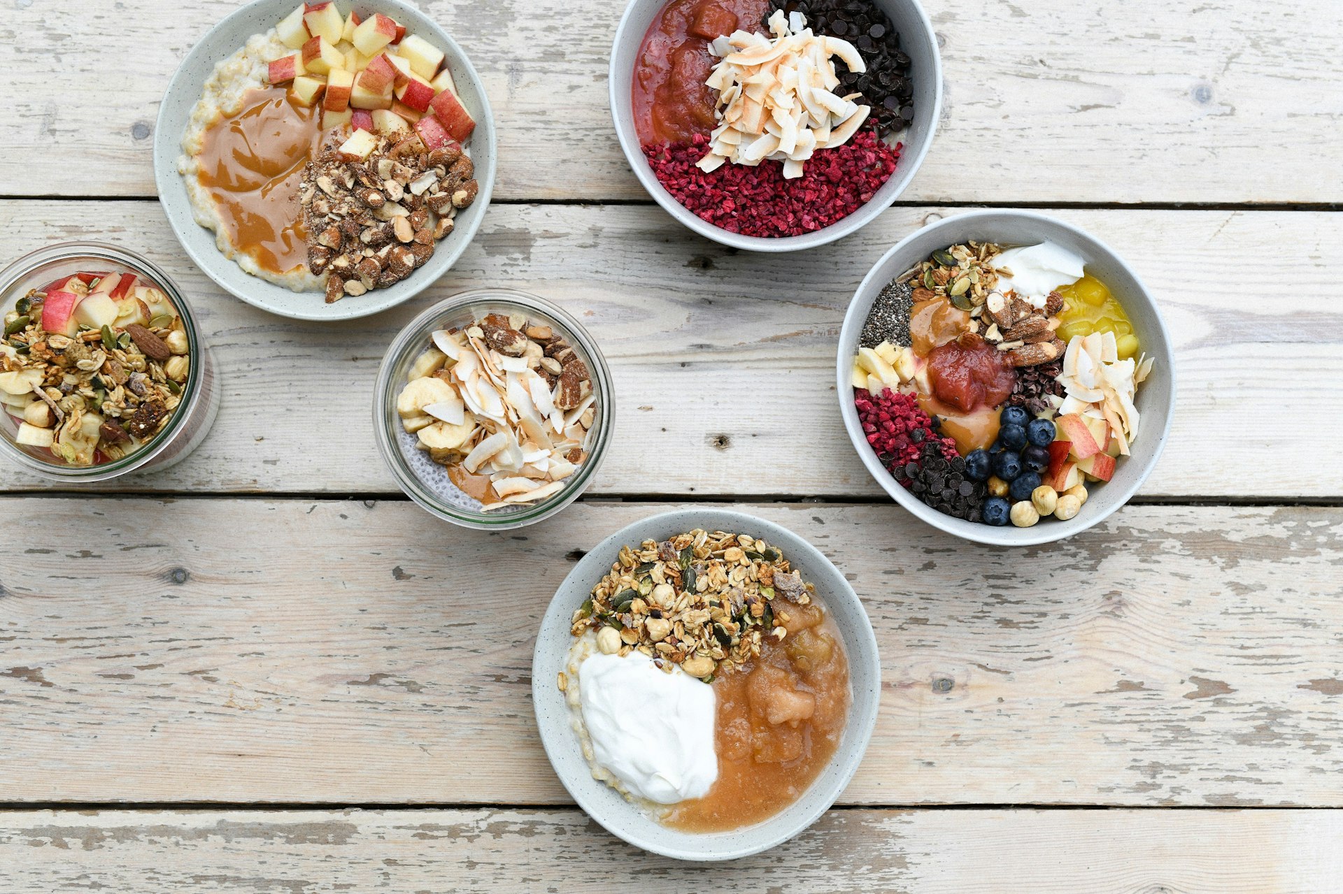 Four bowls and two glass jars sit on a white wooden table; each is full of different dishes, all being assortments of fruit and nuts.