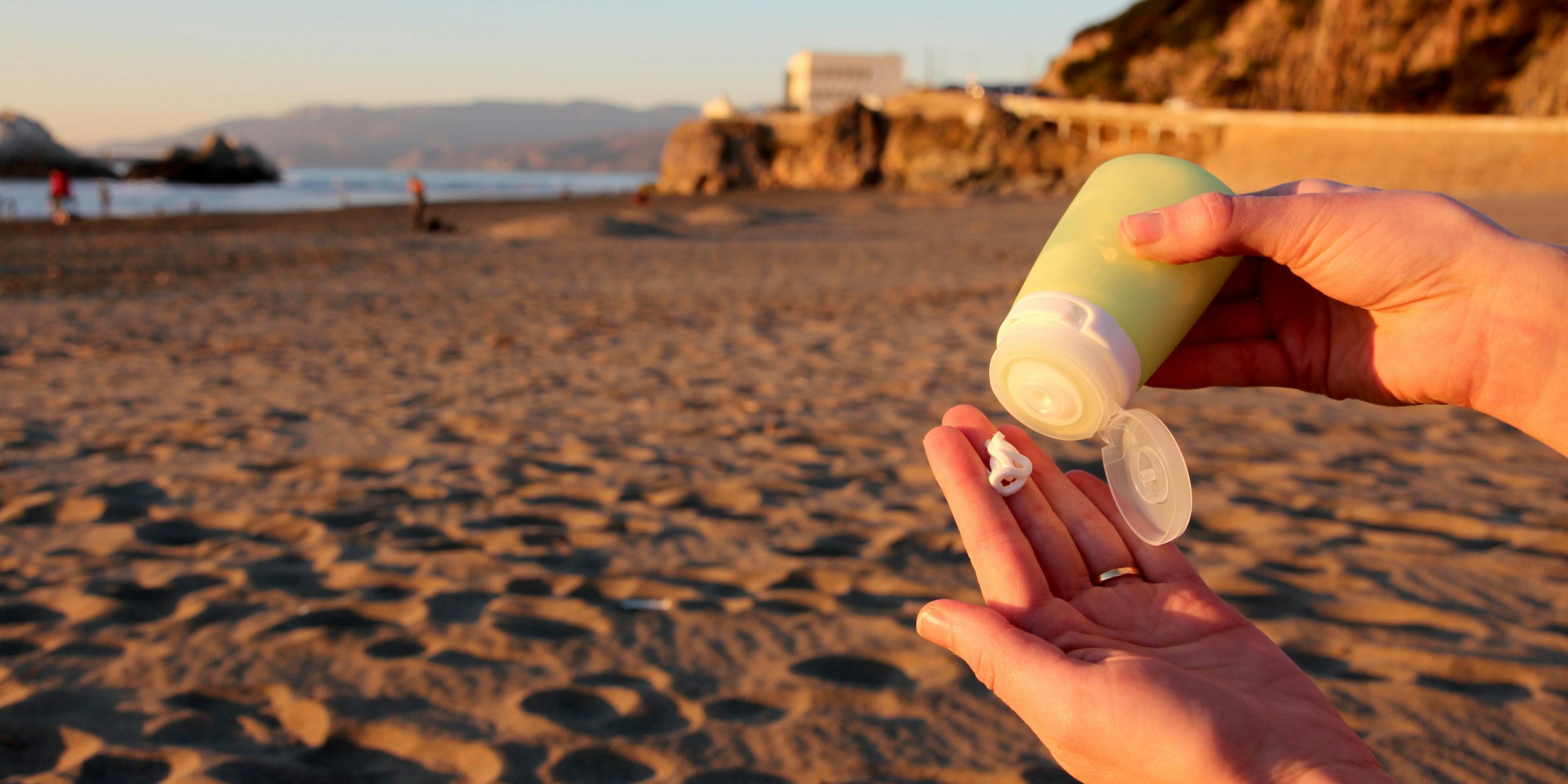 A pair of caucasian hands wearing a gold wedding band squeeze a white cream from a light green humangear GoToob+ on a beach where an out of focus rocky promontory with a white building on it sits in the distance and other beachgoers in red jackets can be seen