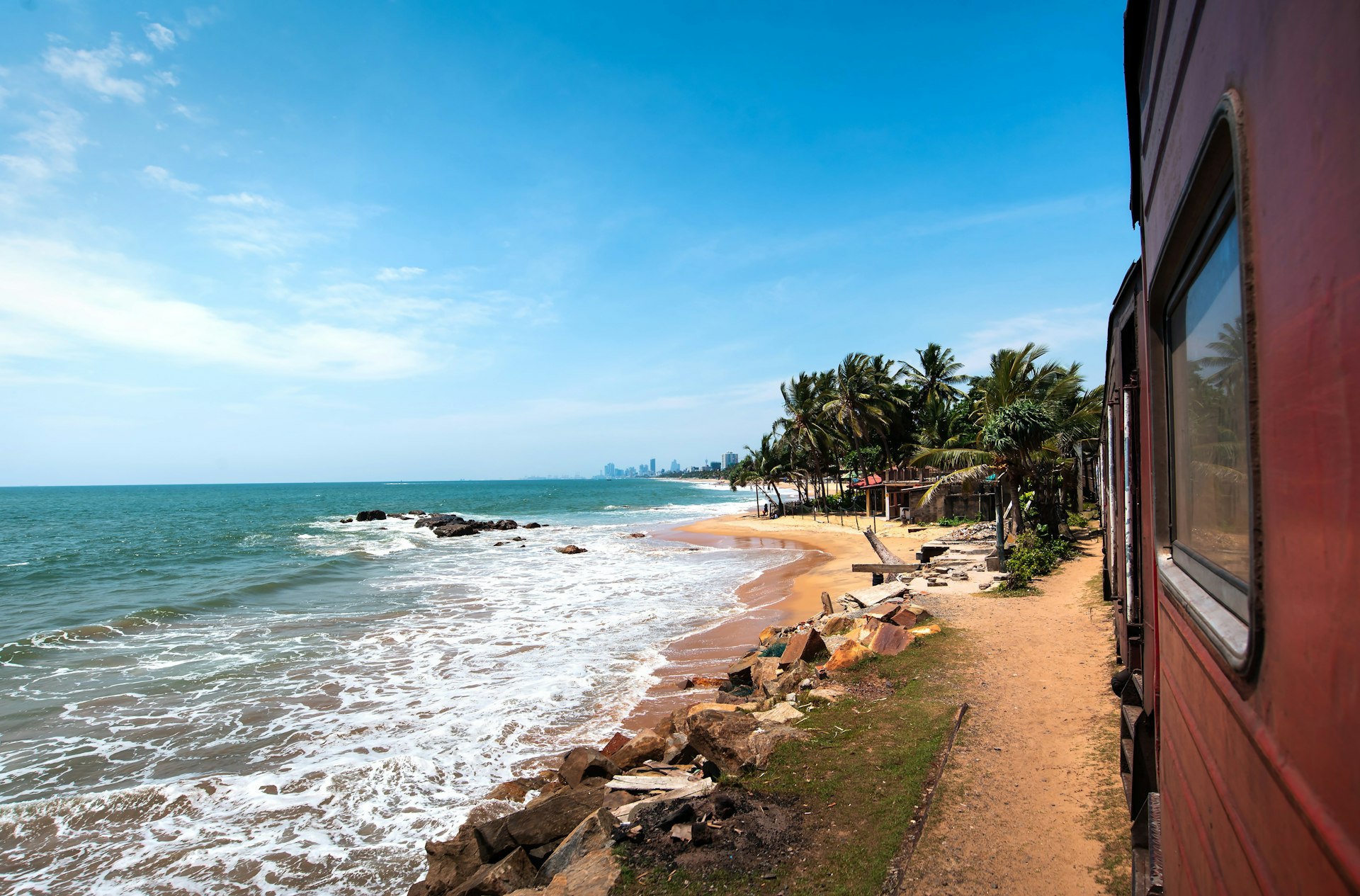 A parallel view of a red train travelling from Colombo to Galle, taken by a passenger on board the train. The tracks runs alongside the coast, with the sea coming right up to the tracks. A small, palm tree-backed beach is visible in the middle ground, while in the distance, the skyscrapers of a metropolis, presumably Colombo, can be seen.
