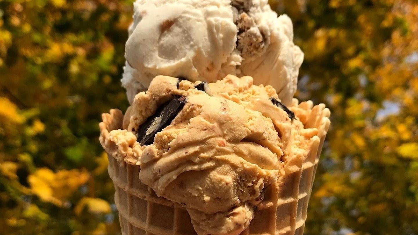 A waffle cone filled with two scoops of ice cream is held up against a backdrop of autumn leaves.