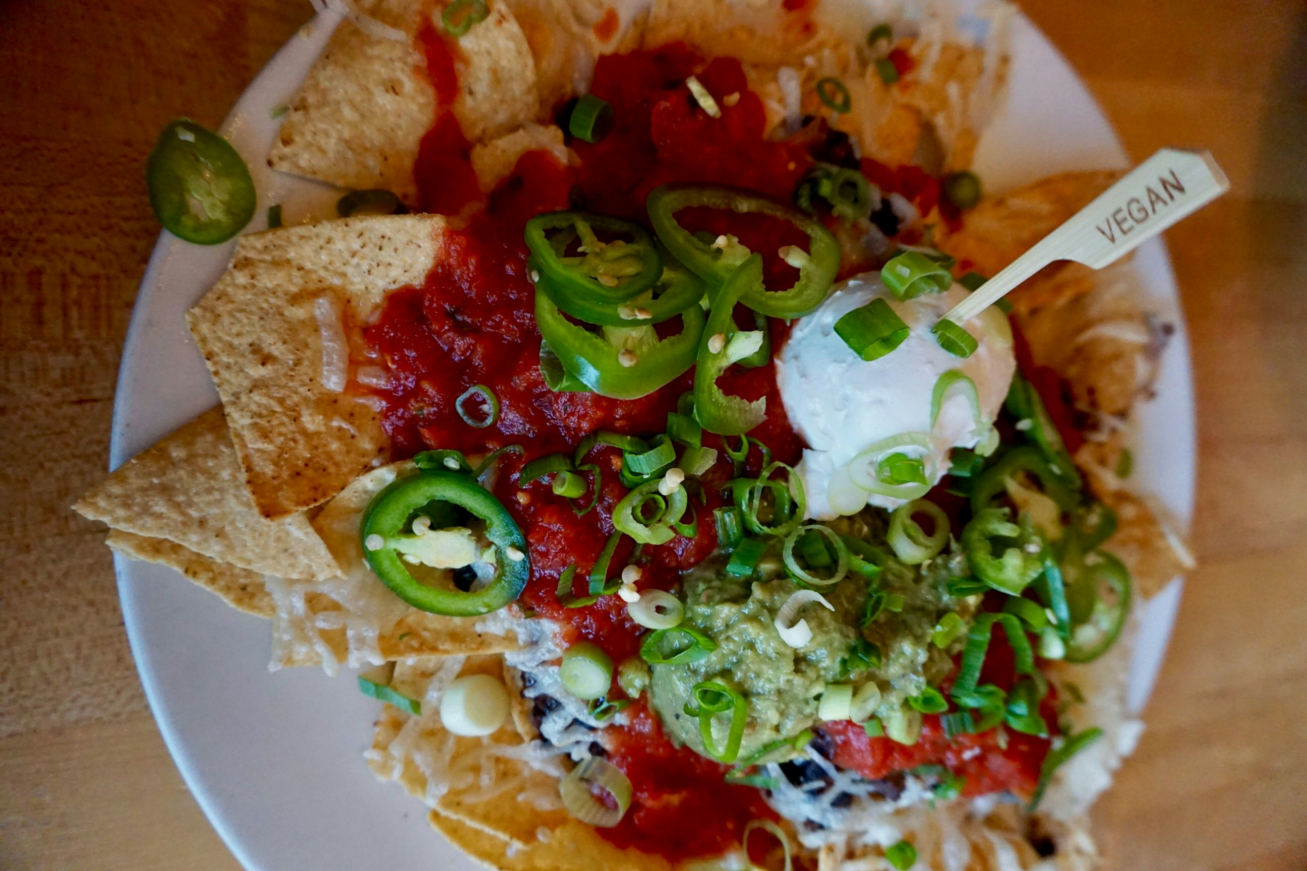 A plate of vegan nachos, piled high with tortilla chips, bright red salsa and guacamole and topped with slices of jalapenos, spring onions and a serving of vegan sour cream. There's a  wooden stick labeled 'vegan' sticking out of the cream.