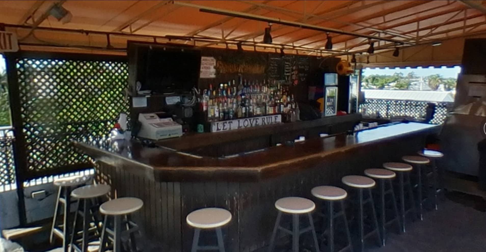 The wood-paneled bar at the Garden of Eden nudist resort is well-worn around the edges, with white stools and shaded by an orange awning. Behind the bar are colorful rows of liquor bottles, and a white shabby-chic sign that reads Let Love Rule