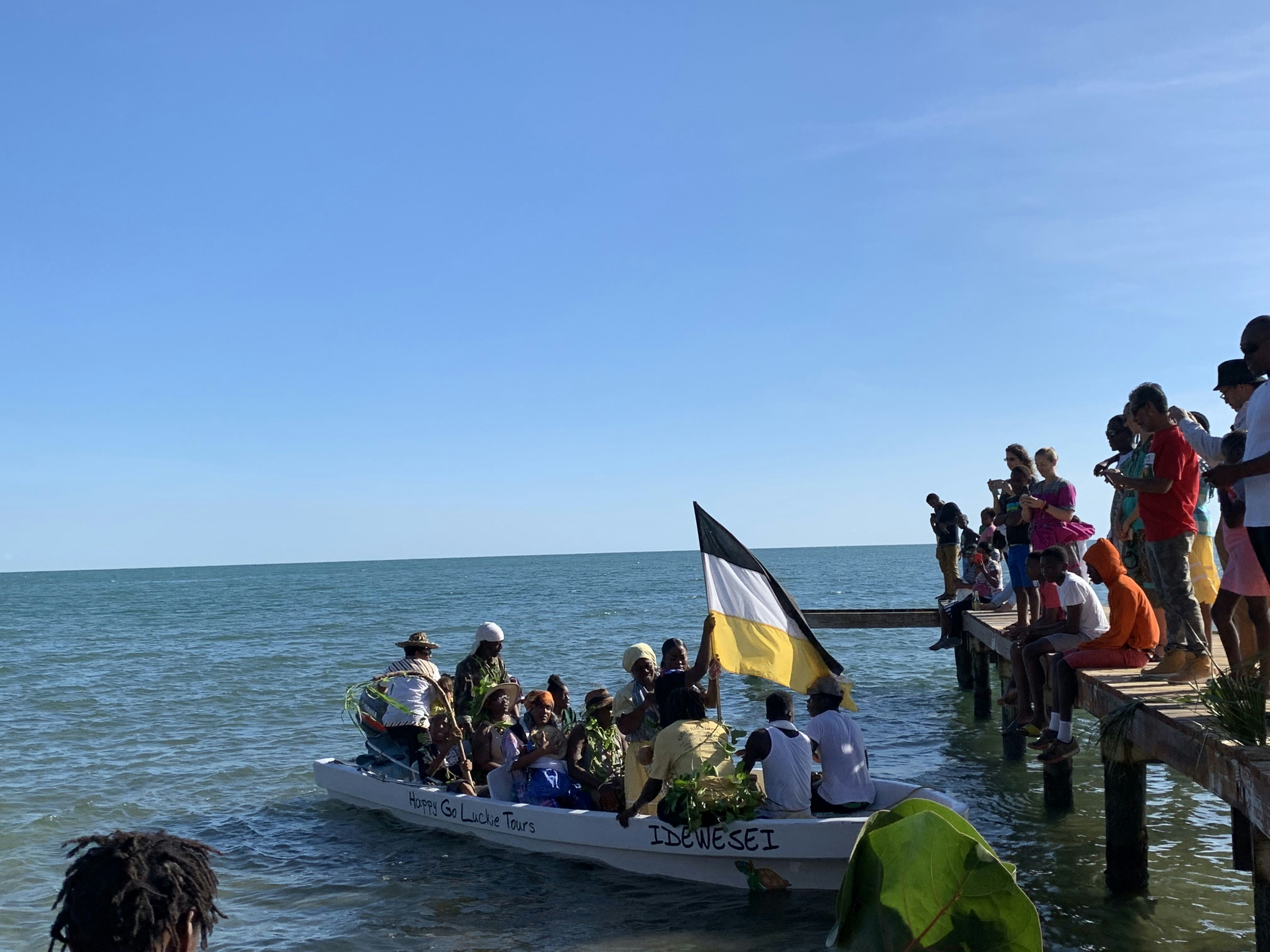A large boat filled with actors re-enact Garifuna Settlement Day as a group of people watch from a wooden pier. 