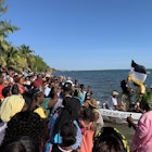 A large group of people stand on the shore watching a pair of boats filled with actors re-enact Garifuna Settle Day.