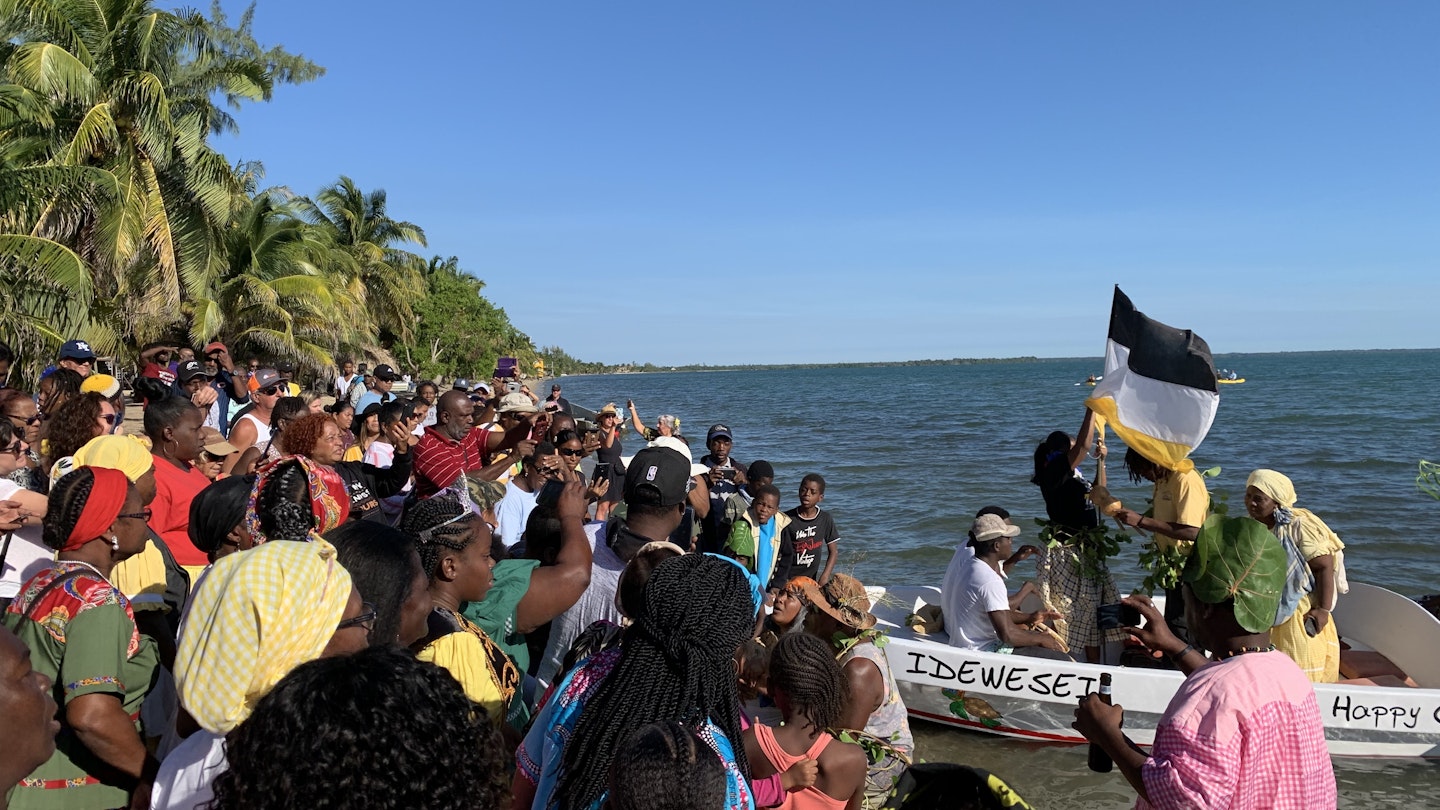 A large group of people stand on the shore watching a pair of boats filled with actors re-enact Garifuna Settle Day.