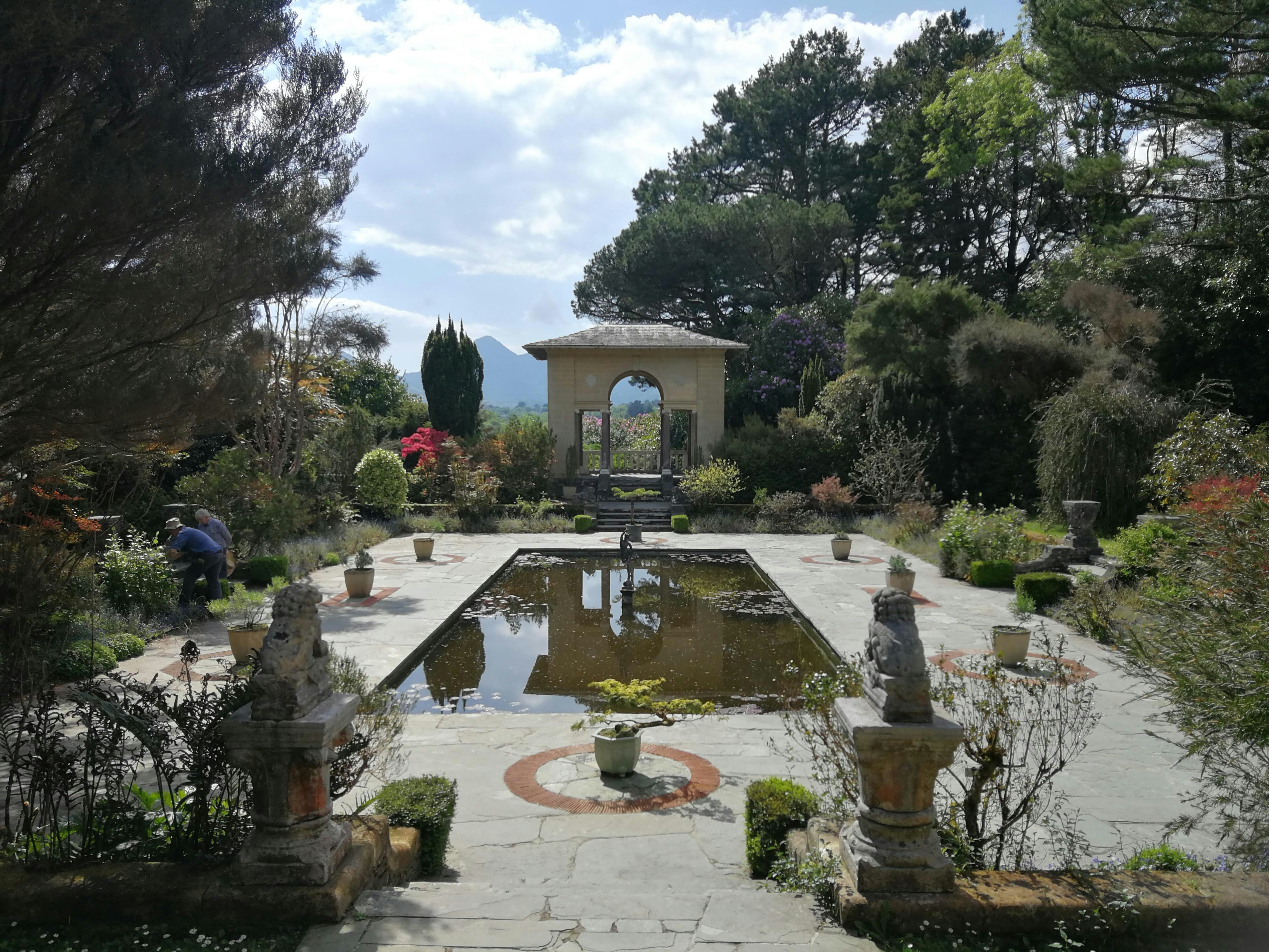 A variety of trees in different shades of green surround a Mediterranean-style terrace that has an attractive sandstone arch at one end, and a rectangular pond stretching towards it.