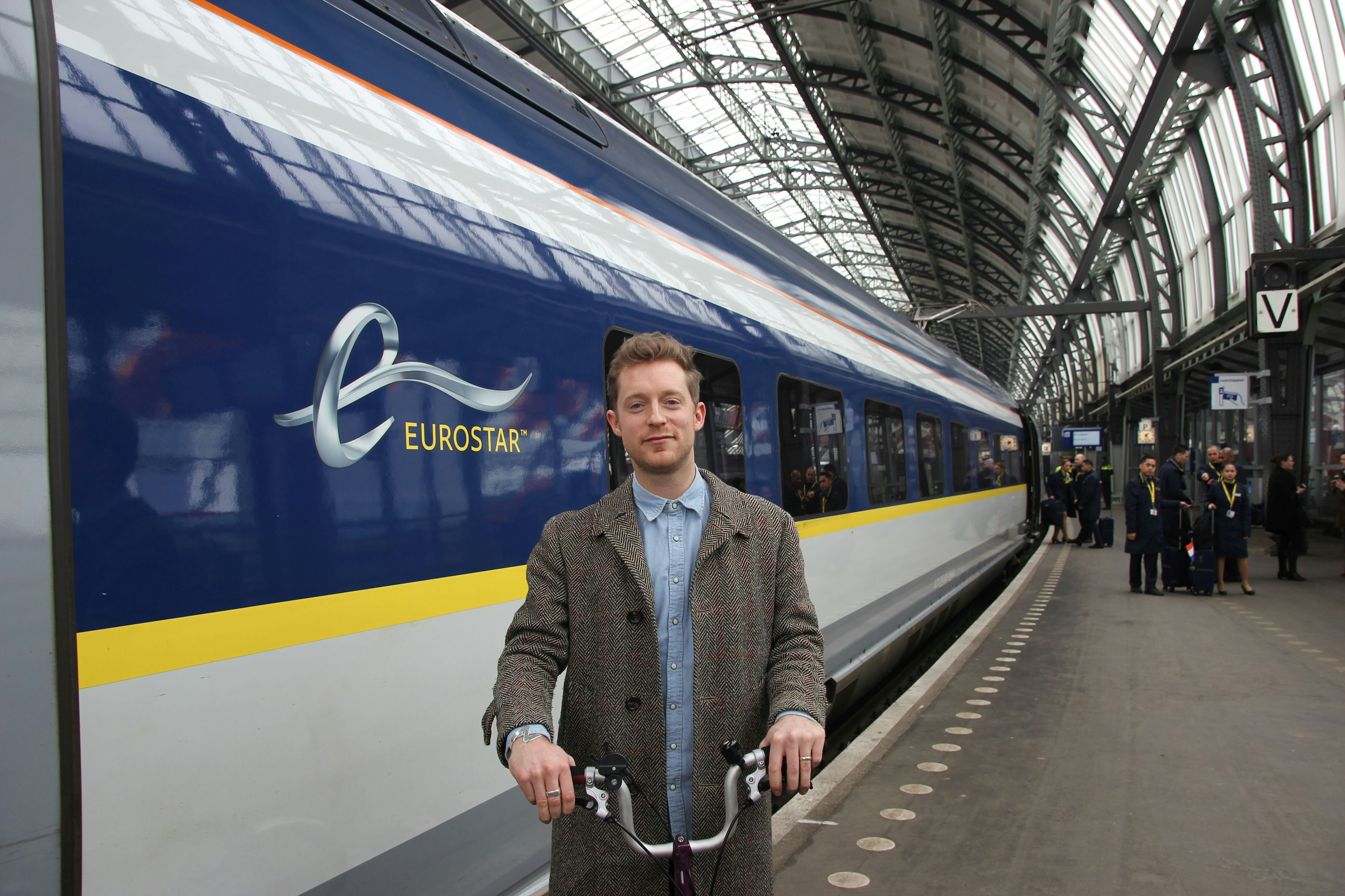 A white man wearing a brown jacket and blue shirt stands with his bike in front of a train