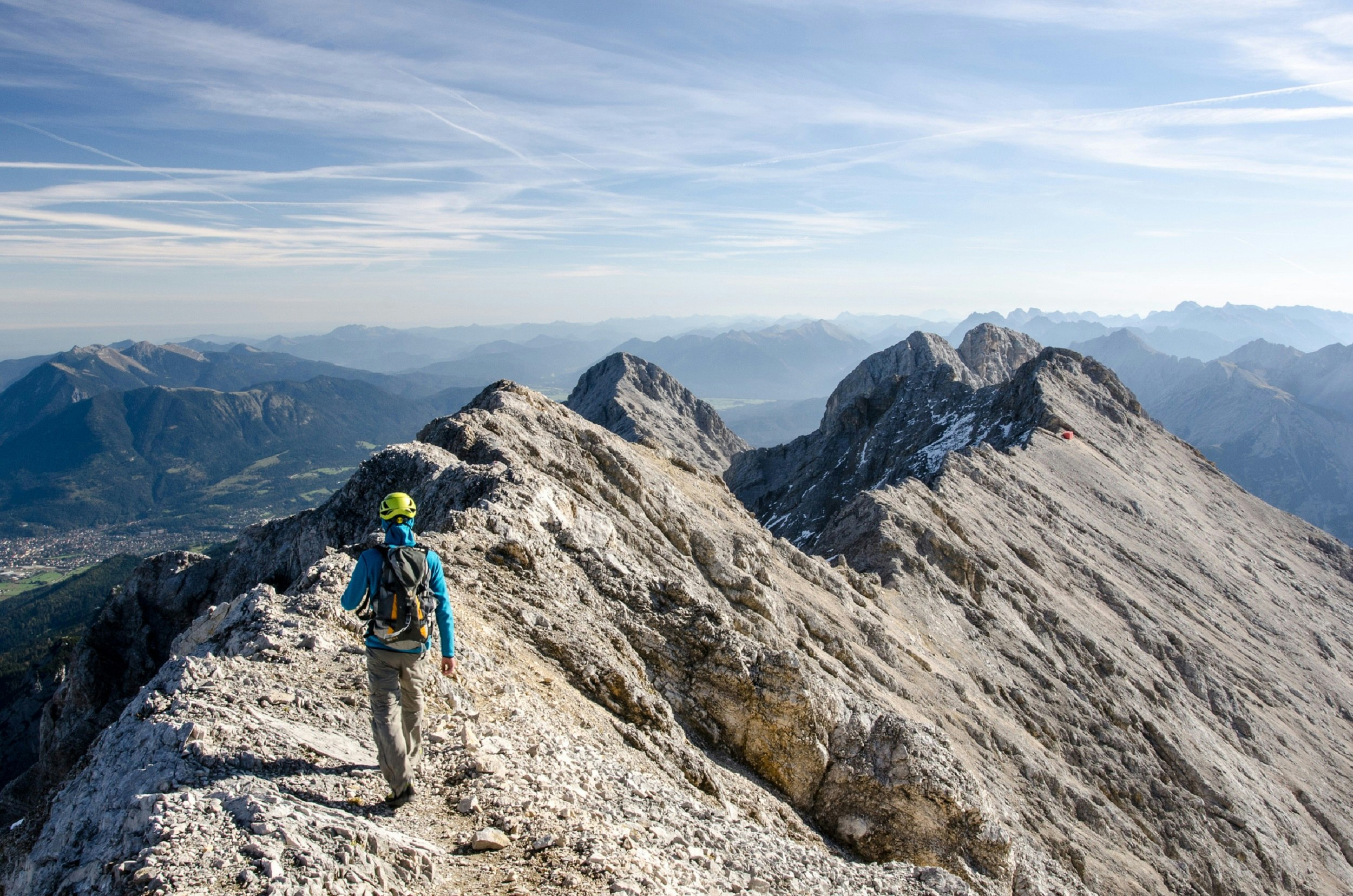 A man in a backpack and helmet walks along a rocky summit ridge high above distant alpine valleys.
