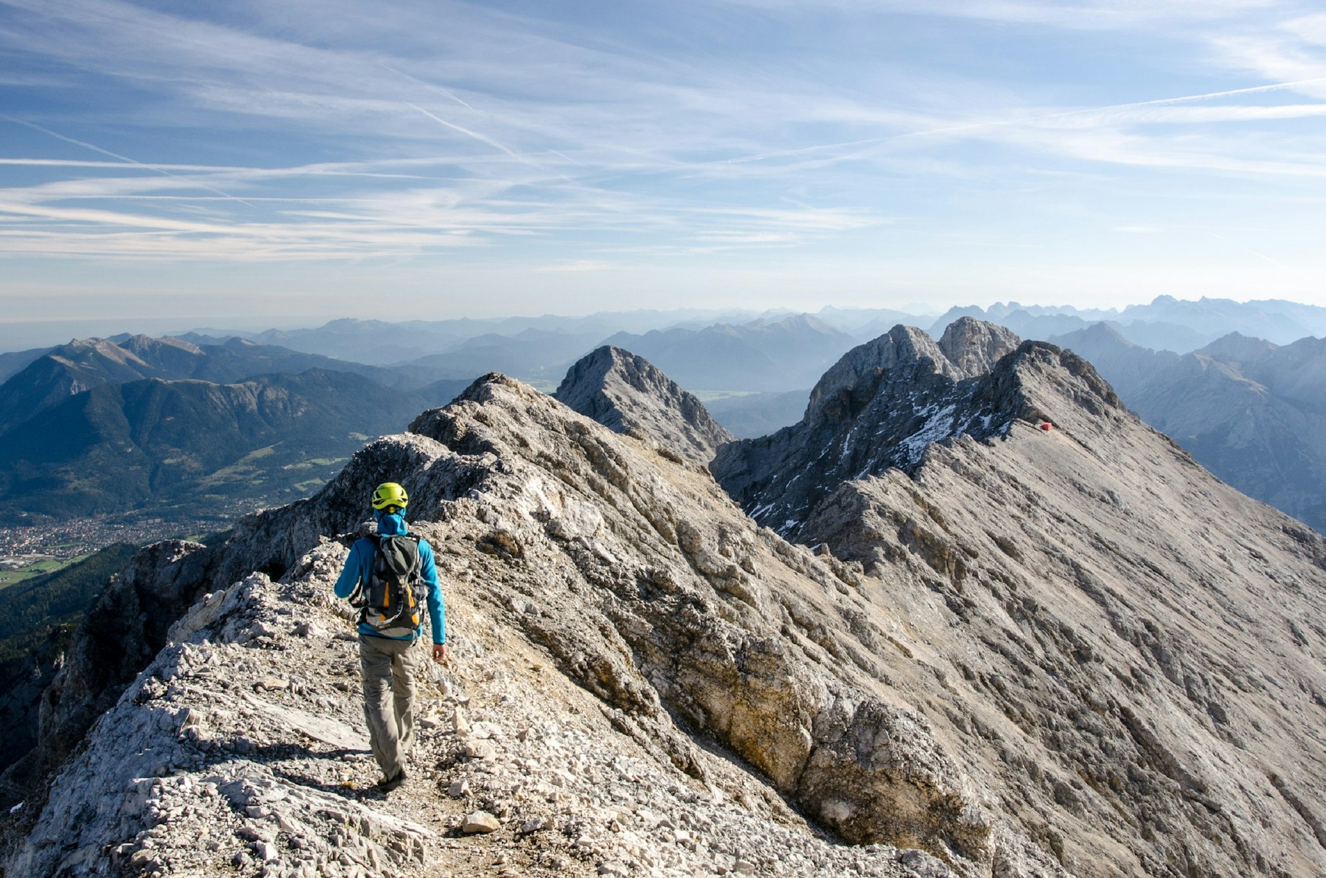 A man in a backpack and helmet walks along a rocky summit ridge high above distant alpine valleys.