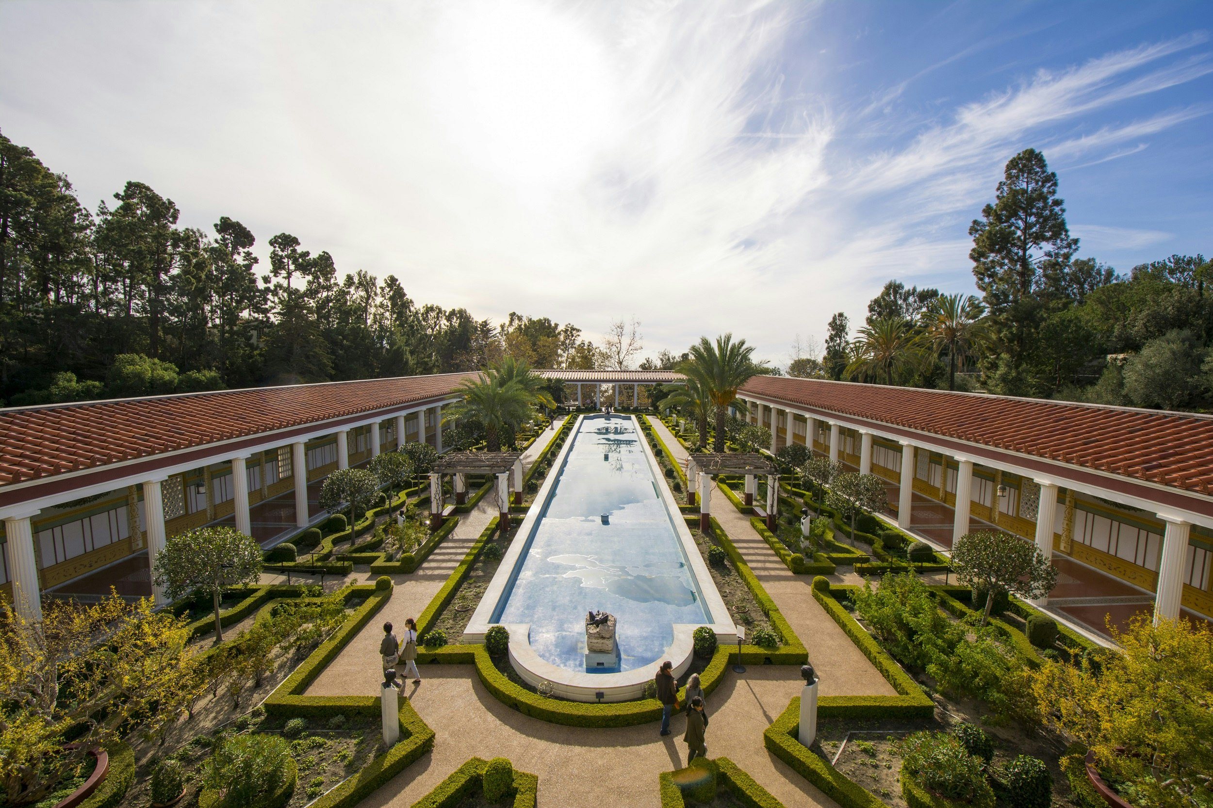 A raised, rectangular garden pond extends into the distance within a massive interior courtyard of the Getty Villa; surrounding the pond are walking paths and manicured shrubbery.